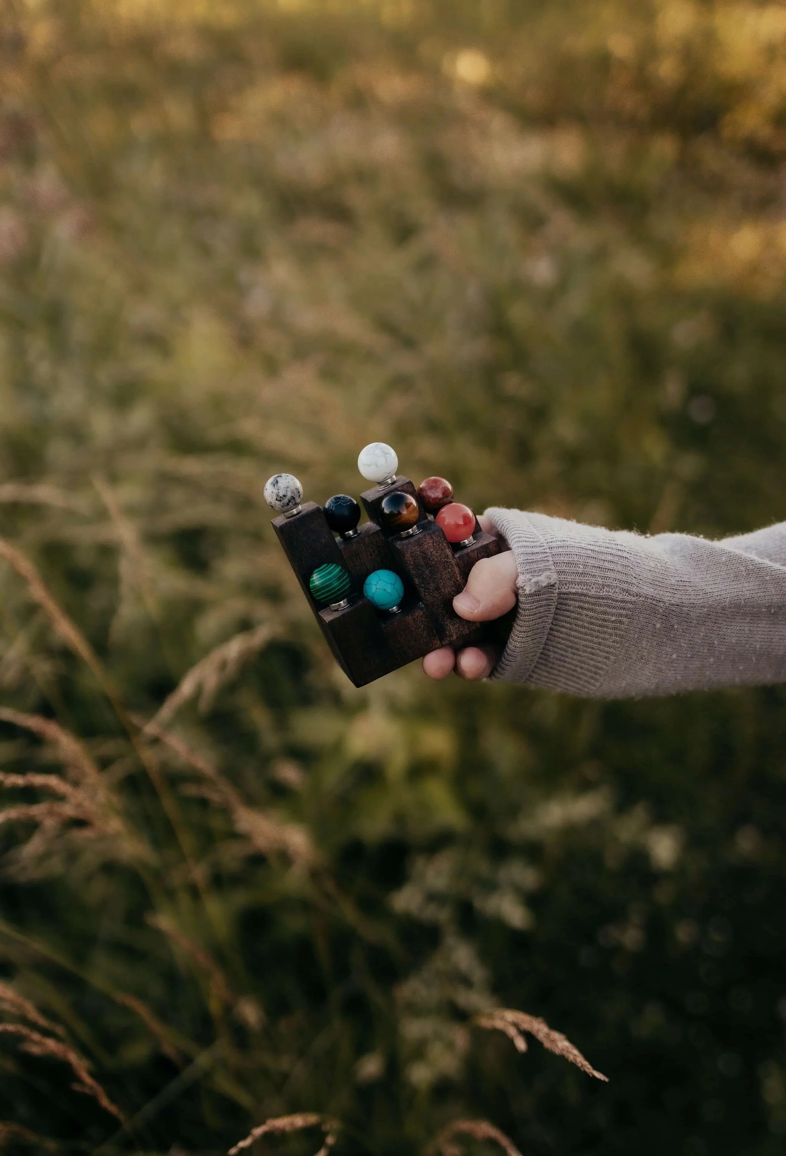 A hand wearing a gray glove holding a black wooden tray with various colored marble balls outdoors, blurred grassy background.