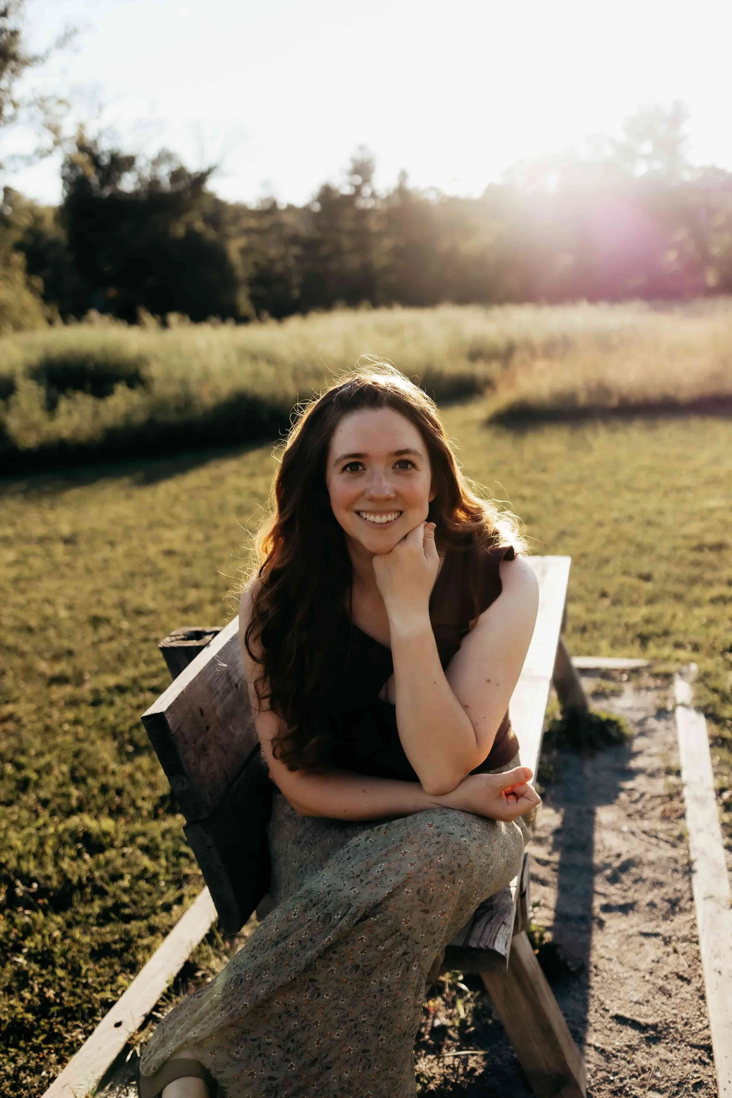 A smiling woman with long wavy hair sitting on a wooden bench outdoors in sunlight, with a grassy field and trees in the background.