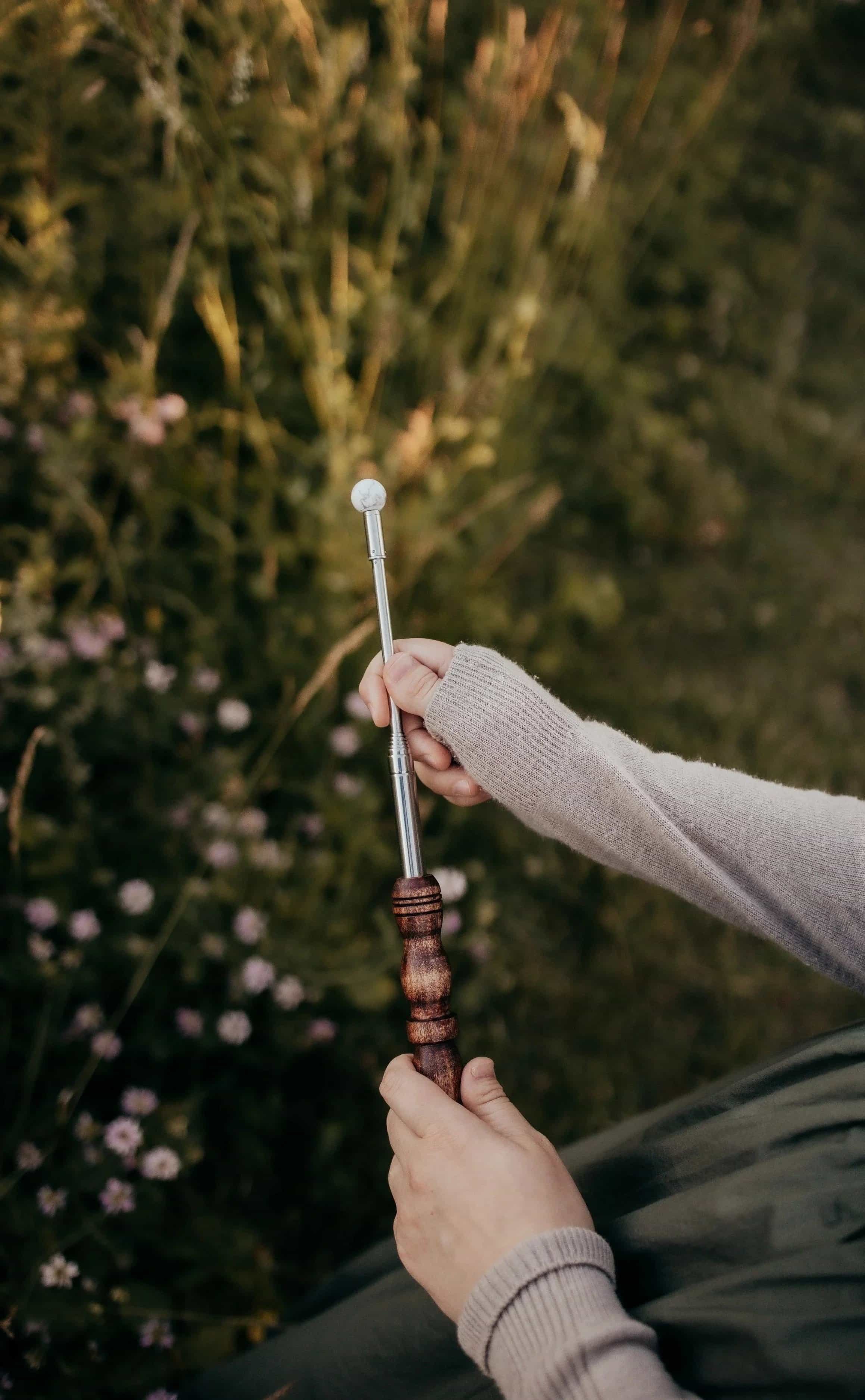 A person holding a metal and wooden gardening tool with a marble ball on top, standing outdoors in front of a background of green bushes and pink flowers.
