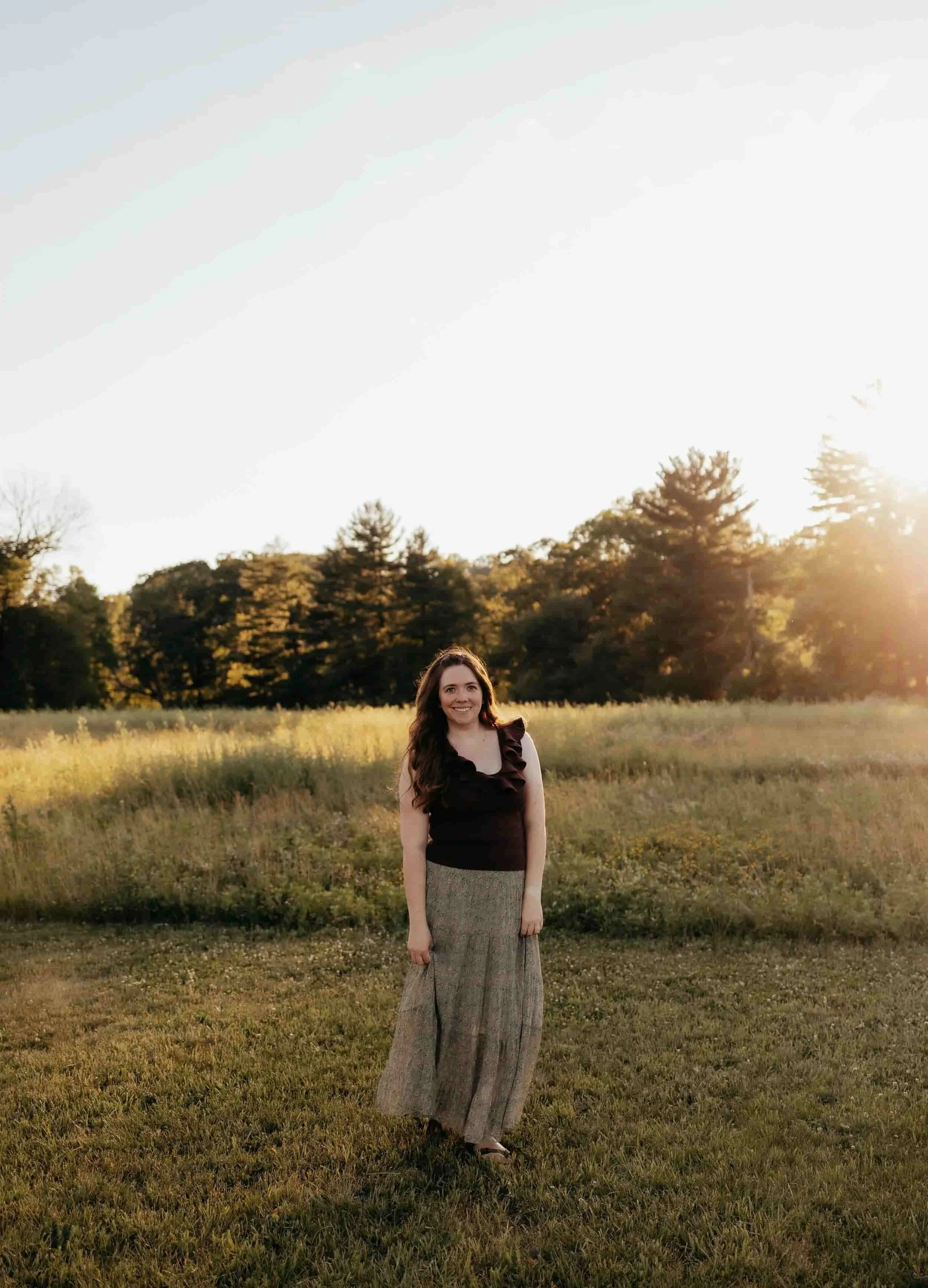 A young woman standing in a grassy field during sunset, with trees in the background and wearing a black top and wide-leg pants.