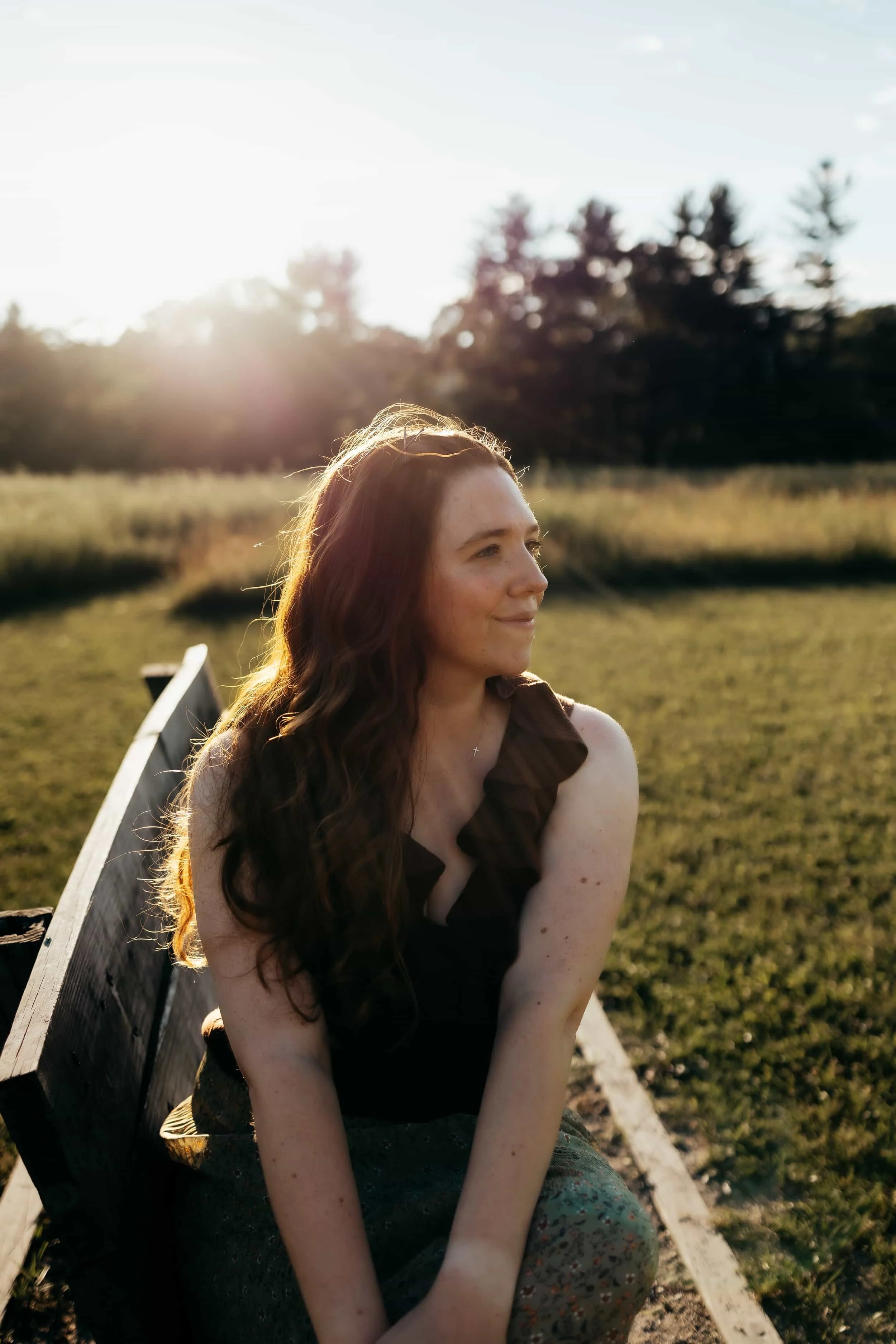 Abigael Davis, LCSW, Christian trauma therapist, sitting on a park bench, smiling while looking to the side, with sunlight shining behind her and trees in the background.