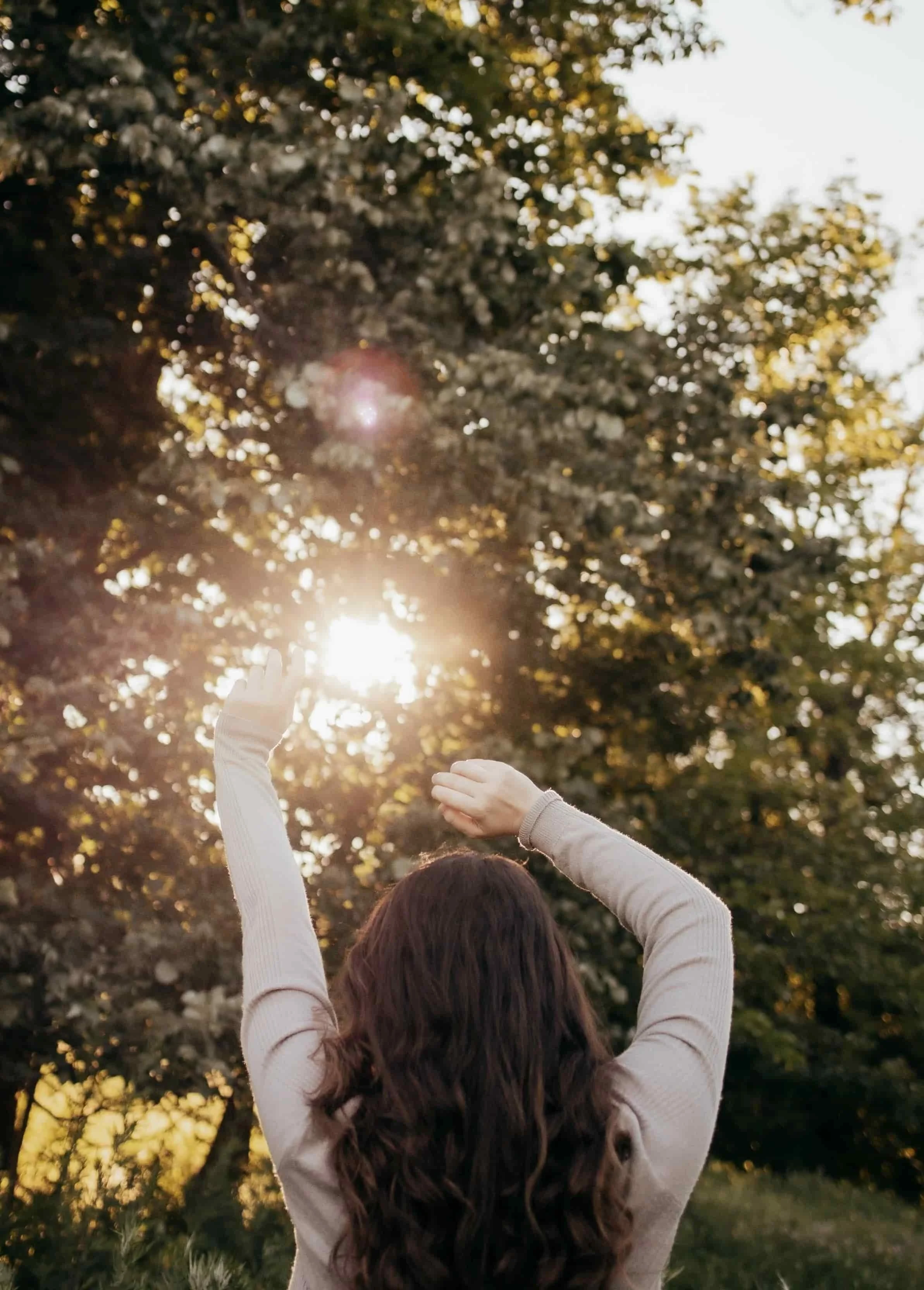 A woman with long brown hair reaching towards the sky as the sun shines through leafy trees during a sunny day.
