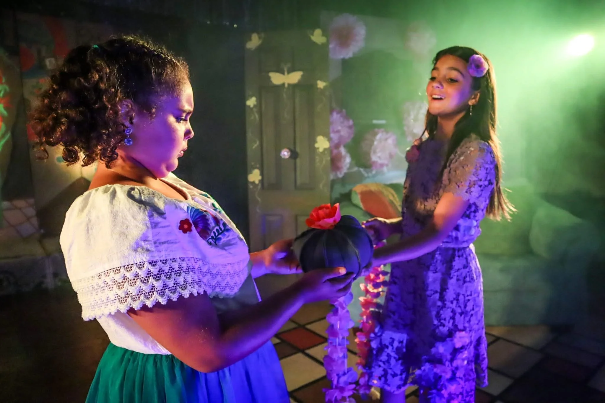 Two young girls exchanging a flower and gift in a colorful, outdoor setting at night, decorated with butterfly and flower motifs.