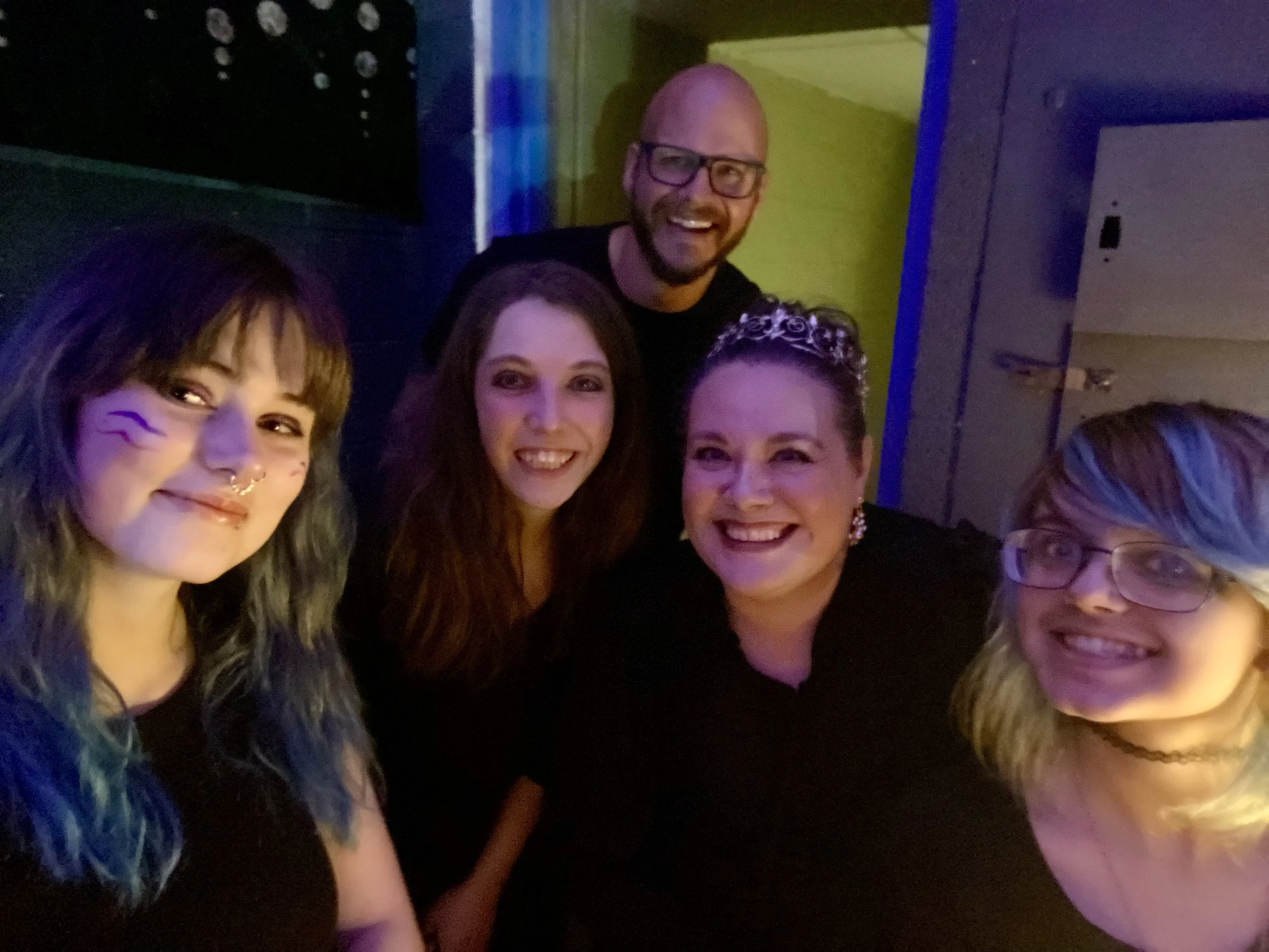Group of five smiling women and one smiling man in a dimly lit room, celebrating or taking a selfie together.