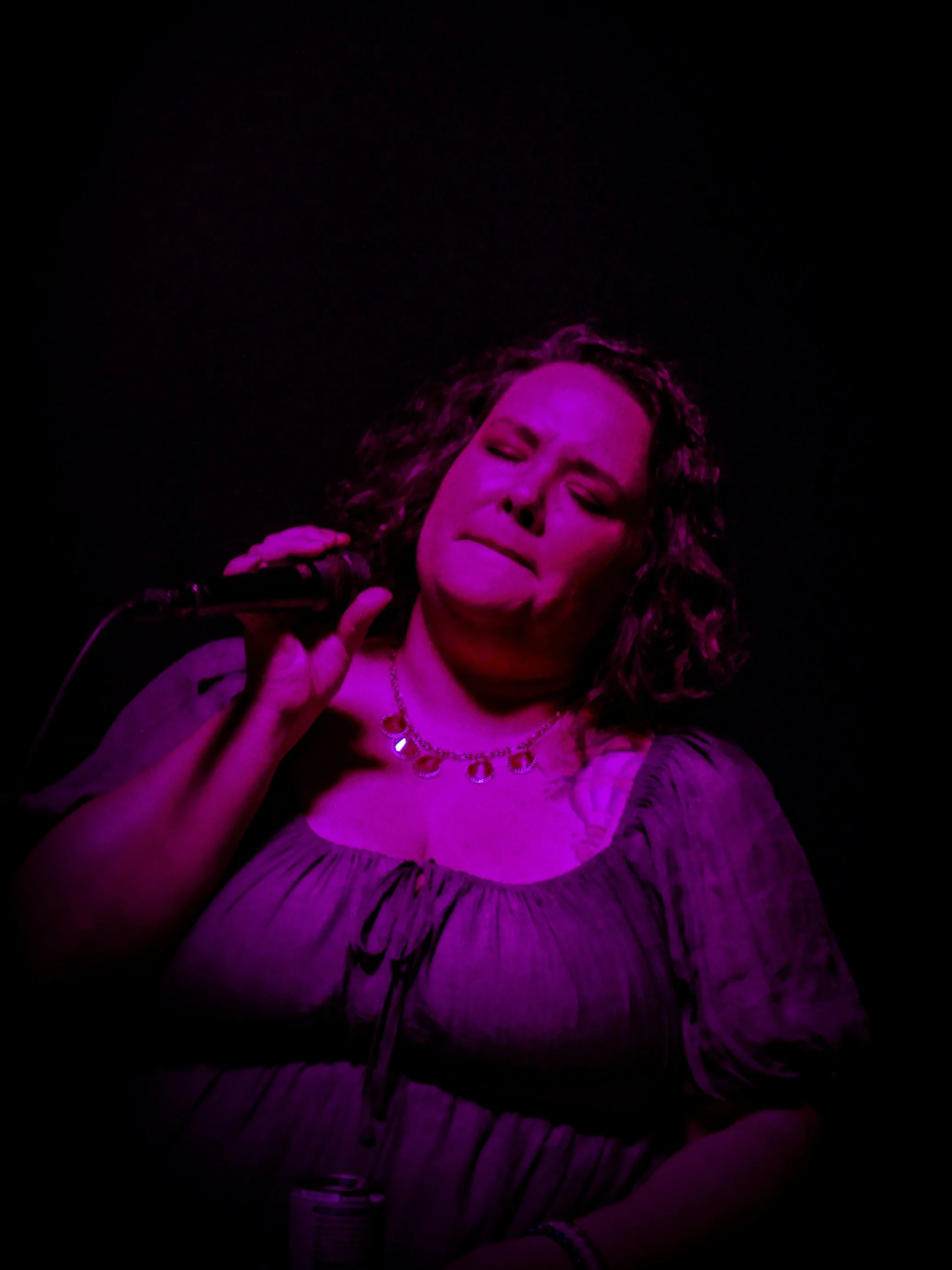 A woman with curly hair singing into a microphone, wearing a ruffled dress and a necklace, with pink stage lighting.