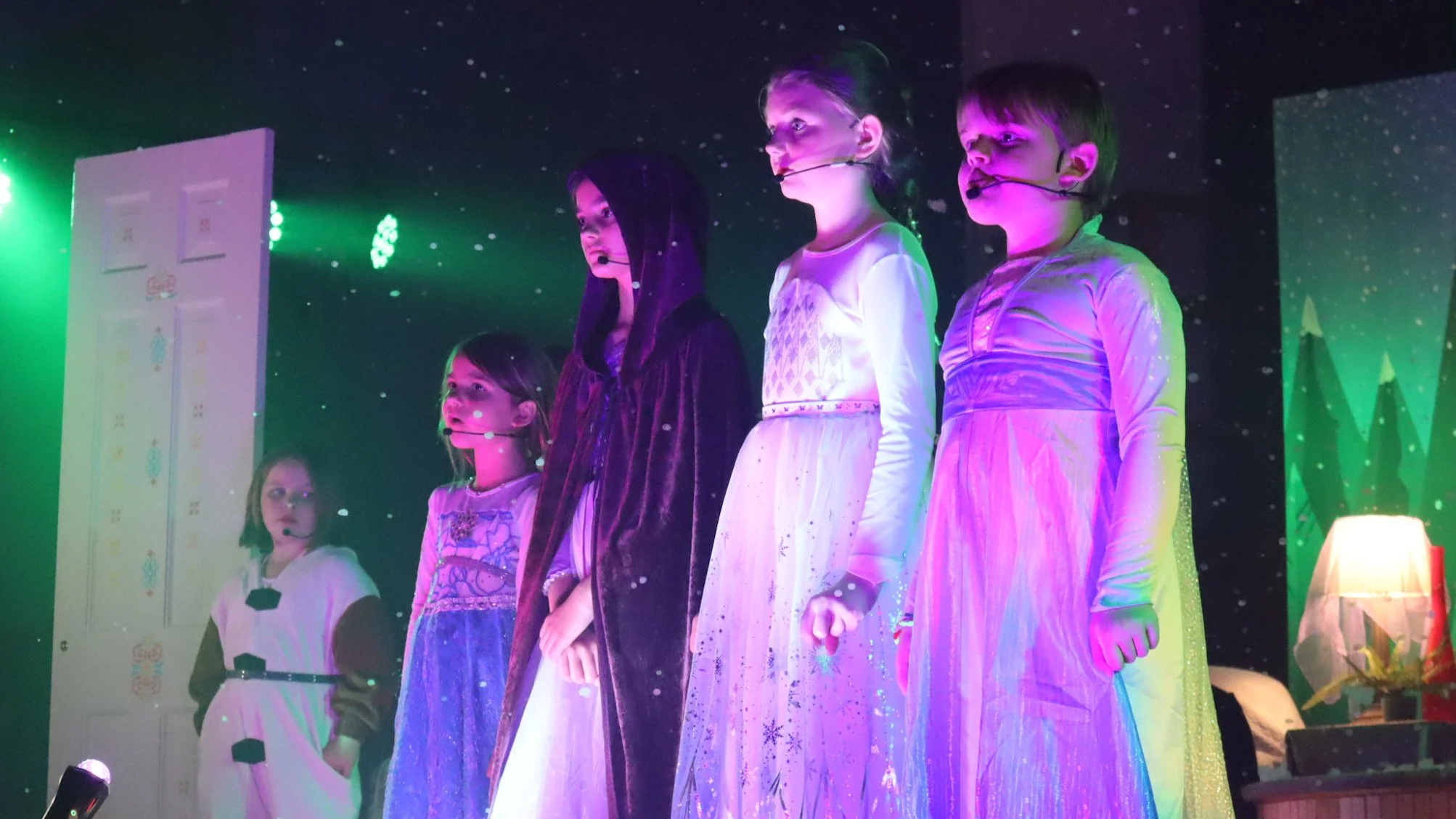 Children dressed in costumes standing on stage under colorful lights, participating in a performance or event.