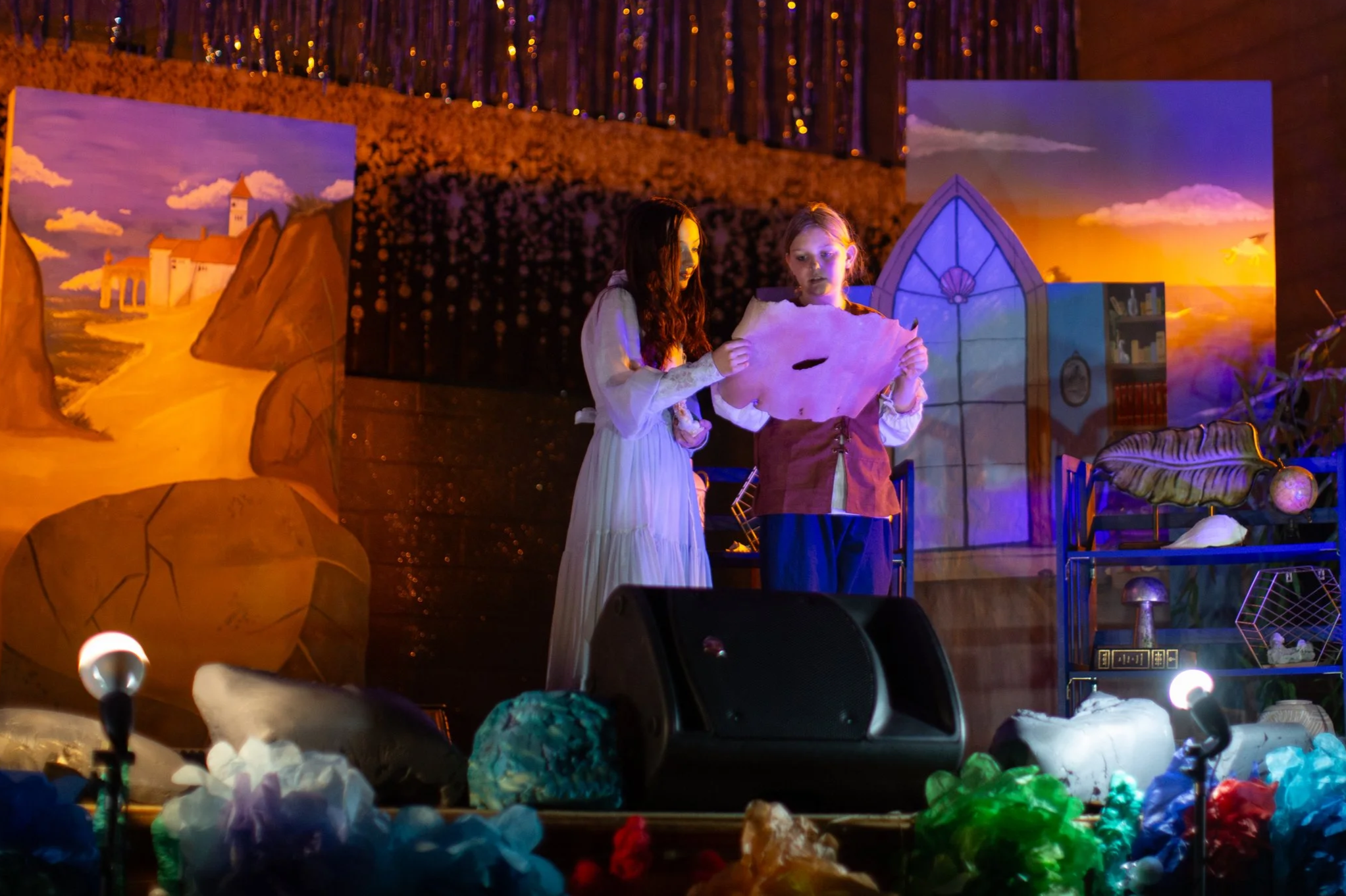 Two young girls dressed as characters from a fairy tale or play, holding a piece of pink paper or cloth, standing on a stage with painted backdrops of a castle and a sunset sky, surrounded by colorful decorations.