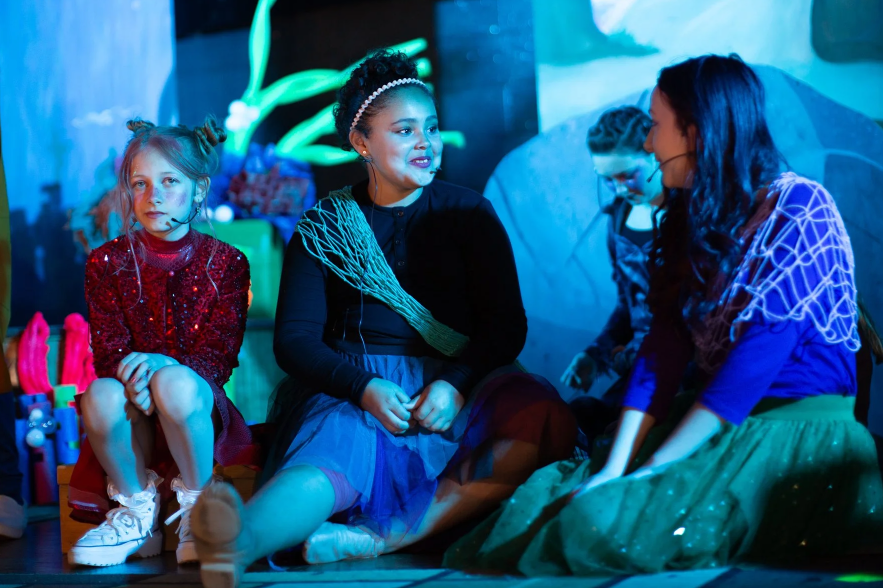 Four young girls sitting on stage, dressed in costumes, engaged in a play or performance, with colorful props and background lighting.