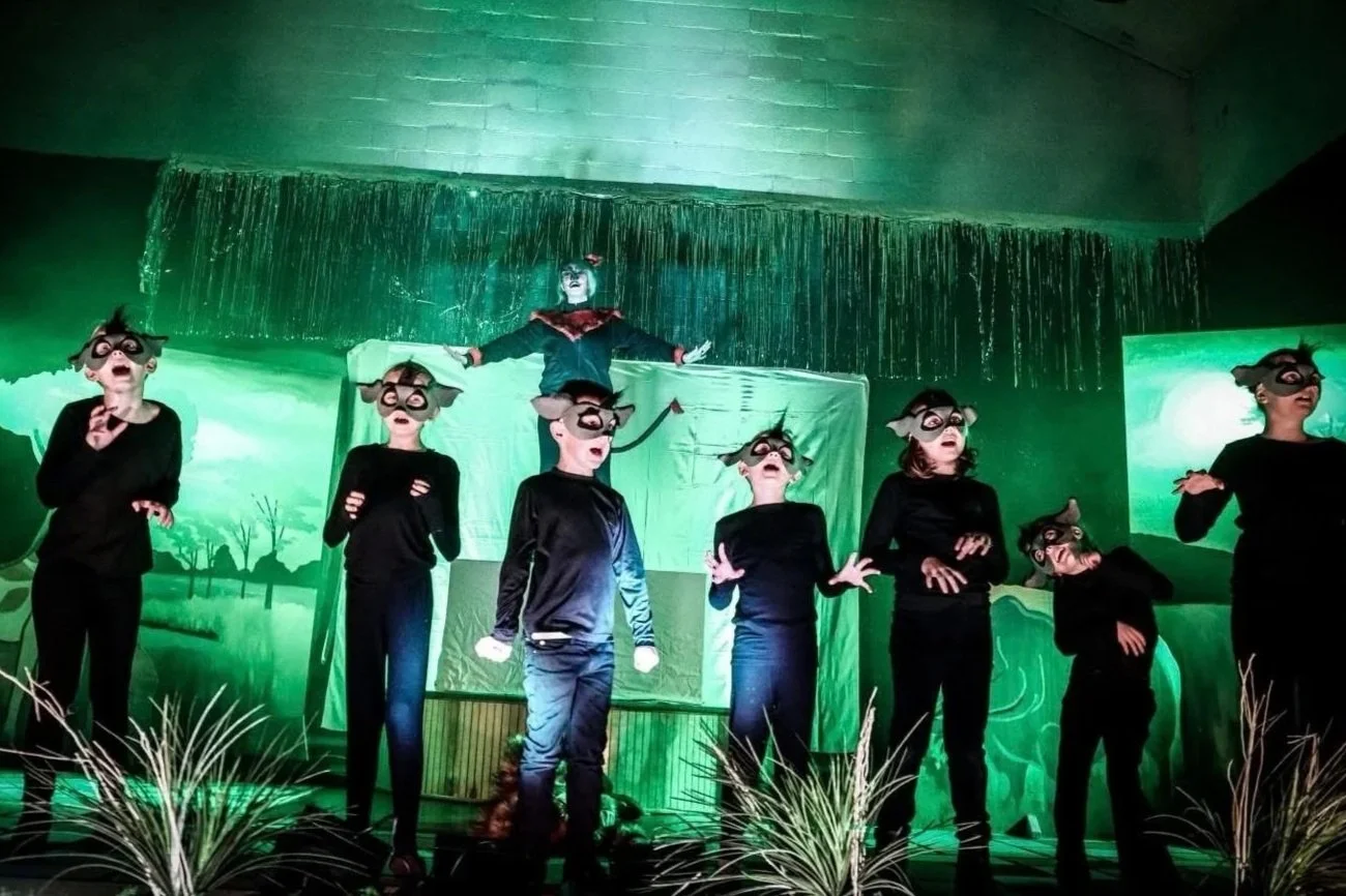 Children on stage wearing animal masks, performing in front of a green scenic backdrop with waterfall and landscape images, with a person elevated above them in theatrical costume.