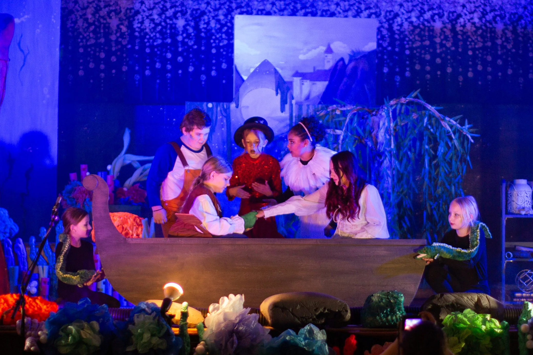Children performing a play on stage with a scenic background, costumes, and props, illuminated by stage lighting.