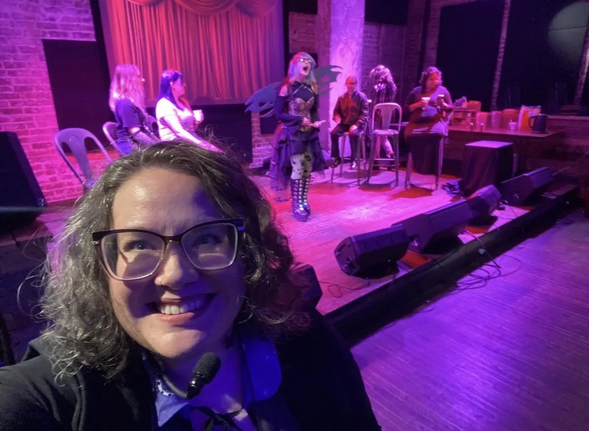 A woman with curly hair and glasses taking a selfie in front of a stage where performers are rehearsing or preparing for a show, with purple and pink stage lighting.