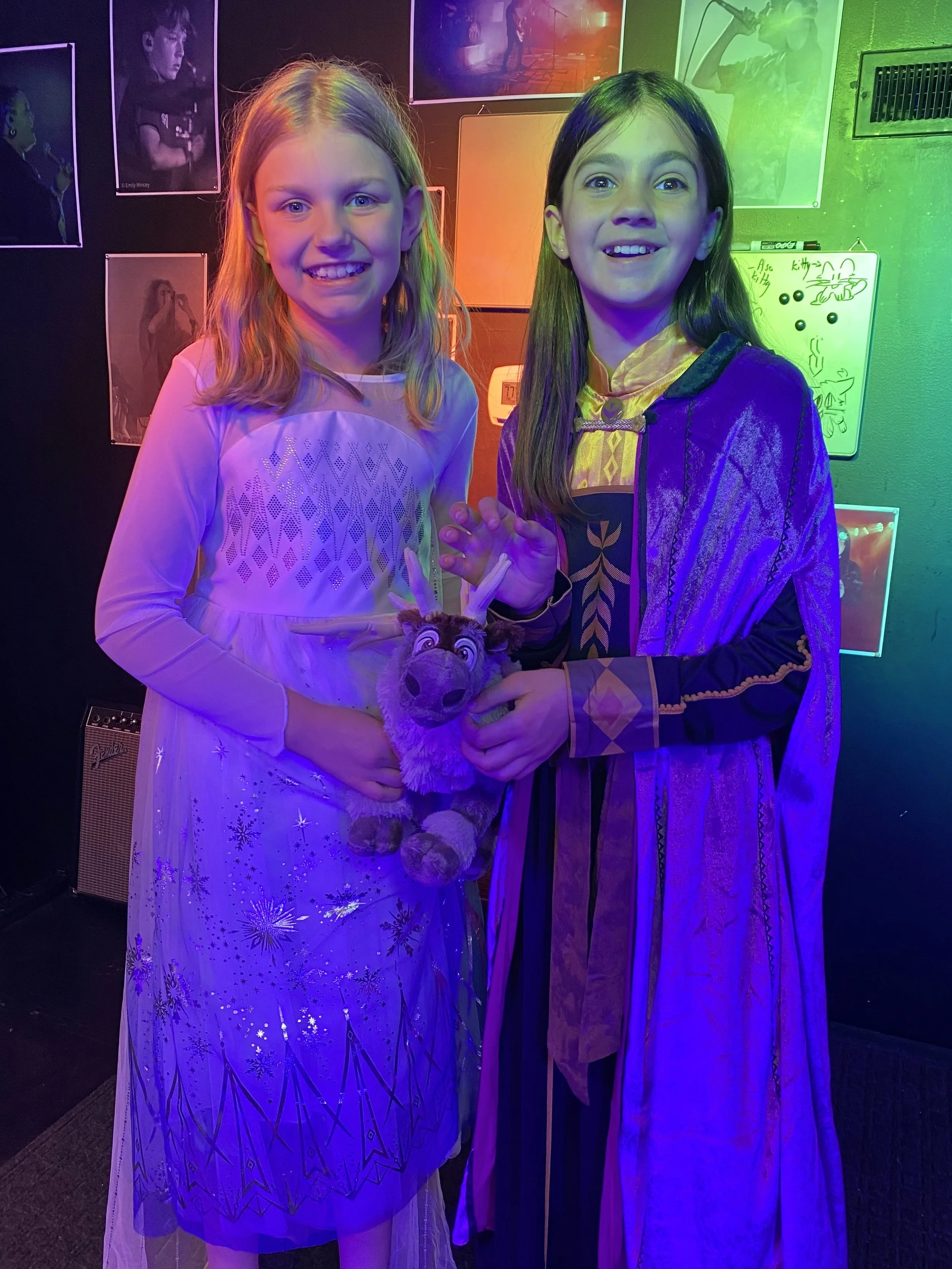 Two young girls dressed in costumes, holding a plush reindeer toy, standing in front of a colorful, illuminated backdrop with artwork and photographs on the wall.