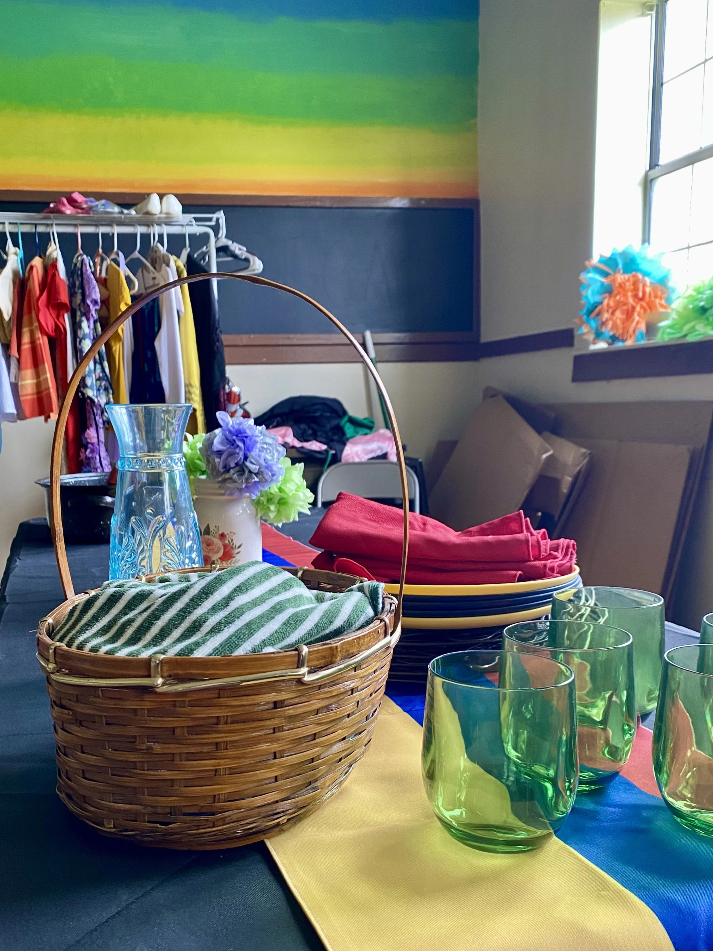 Table with colorful glass cups, a basket with green striped cloth, and a yellow table runner. Behind, tiered trays hold flowers, and a clothing rack displays children's clothing with shoes on top. There is a window with colorful tissue paper decorations and a mural with green, yellow, orange, and blue stripes on the wall.