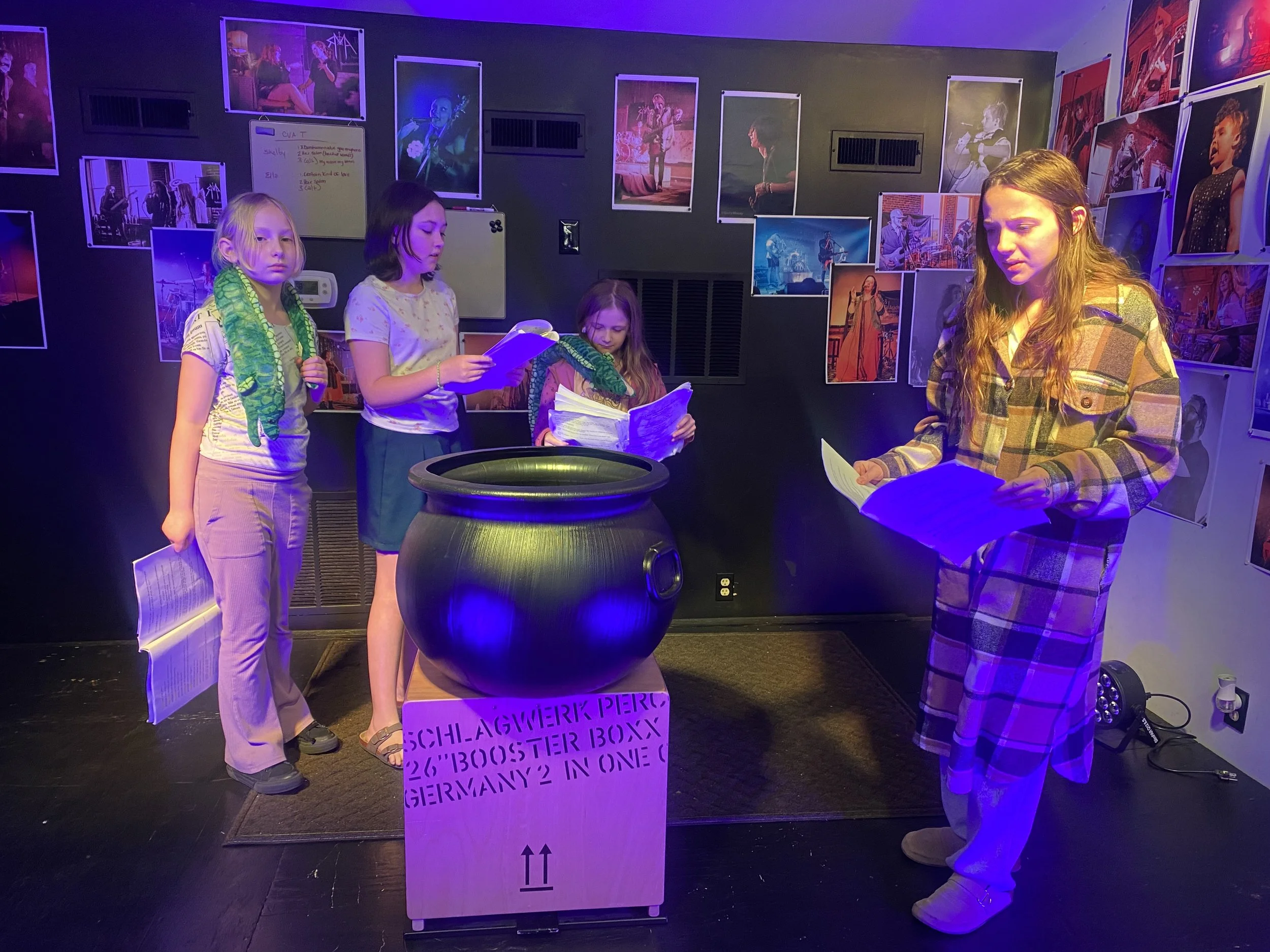 Group of four young girls standing in front of a display with photographs of musicians, holding papers, with a cardboard sign and a large black pot in front. The background is decorated with framed photos of musicians on the wall, under purple lighti
