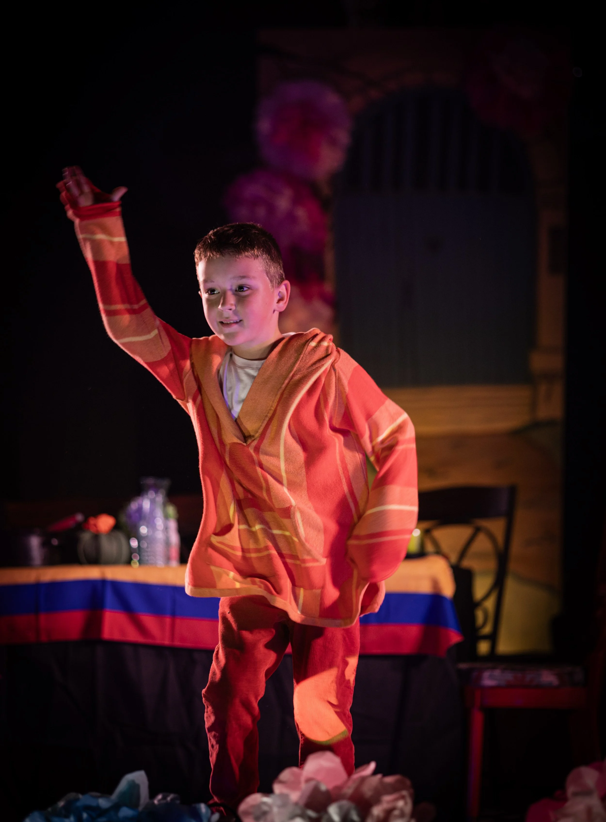A young boy wearing a red and orange patterned hoodie and red pants standing on stage at a Halloween party, with a woman in the background, a table with snacks behind him, and a tablecloth with red, blue, and yellow stripes.