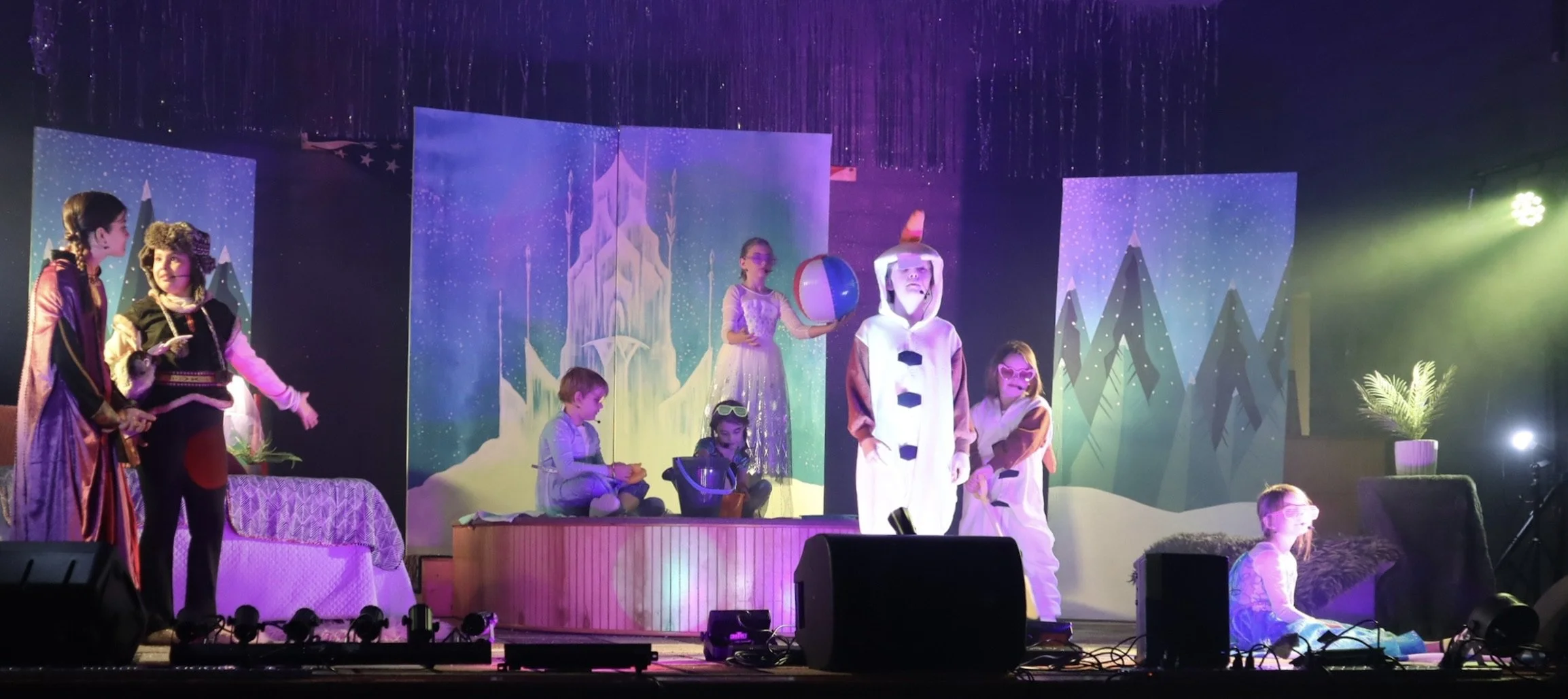 Children performing in a winter-themed stage play, with some dressed as snowmen and others in costumes, set against snow and mountain backdrop with pine trees.