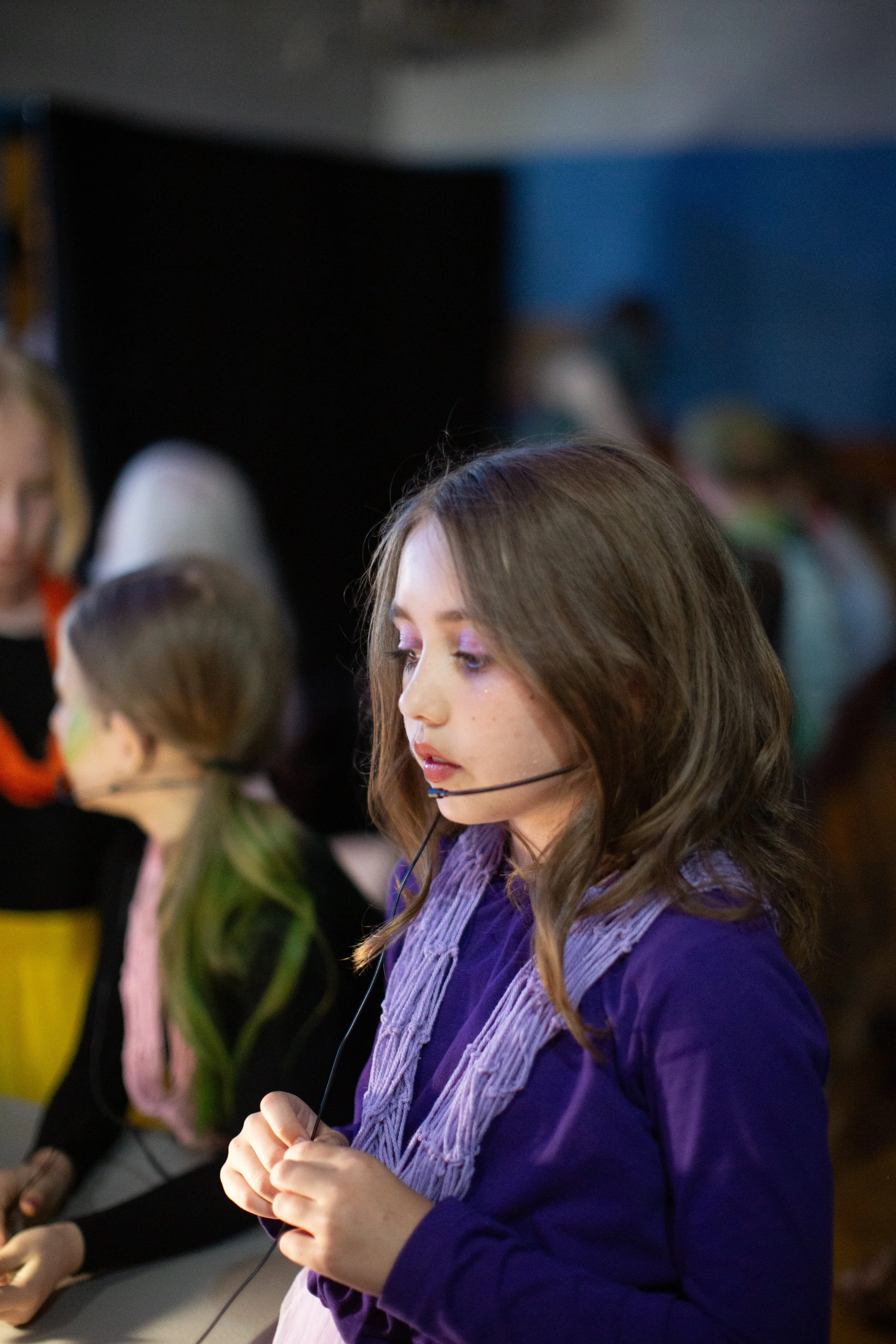 Young woman with long brown hair, wearing a purple shirt and a lavender scarf, holding a microphone headset with her eyes closed in a classroom or seminar setting. Other people are visible blurred in the background.