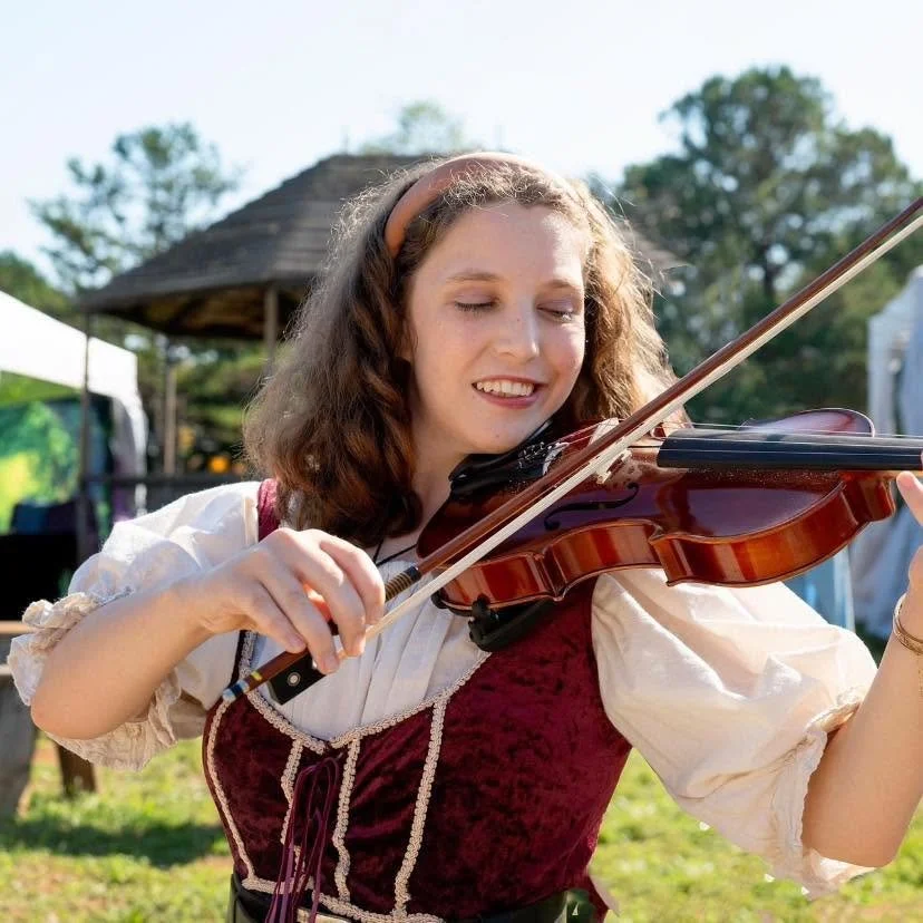 Young woman playing a violin outdoors, wearing a historical or costume dress, with a background of trees and a tent.