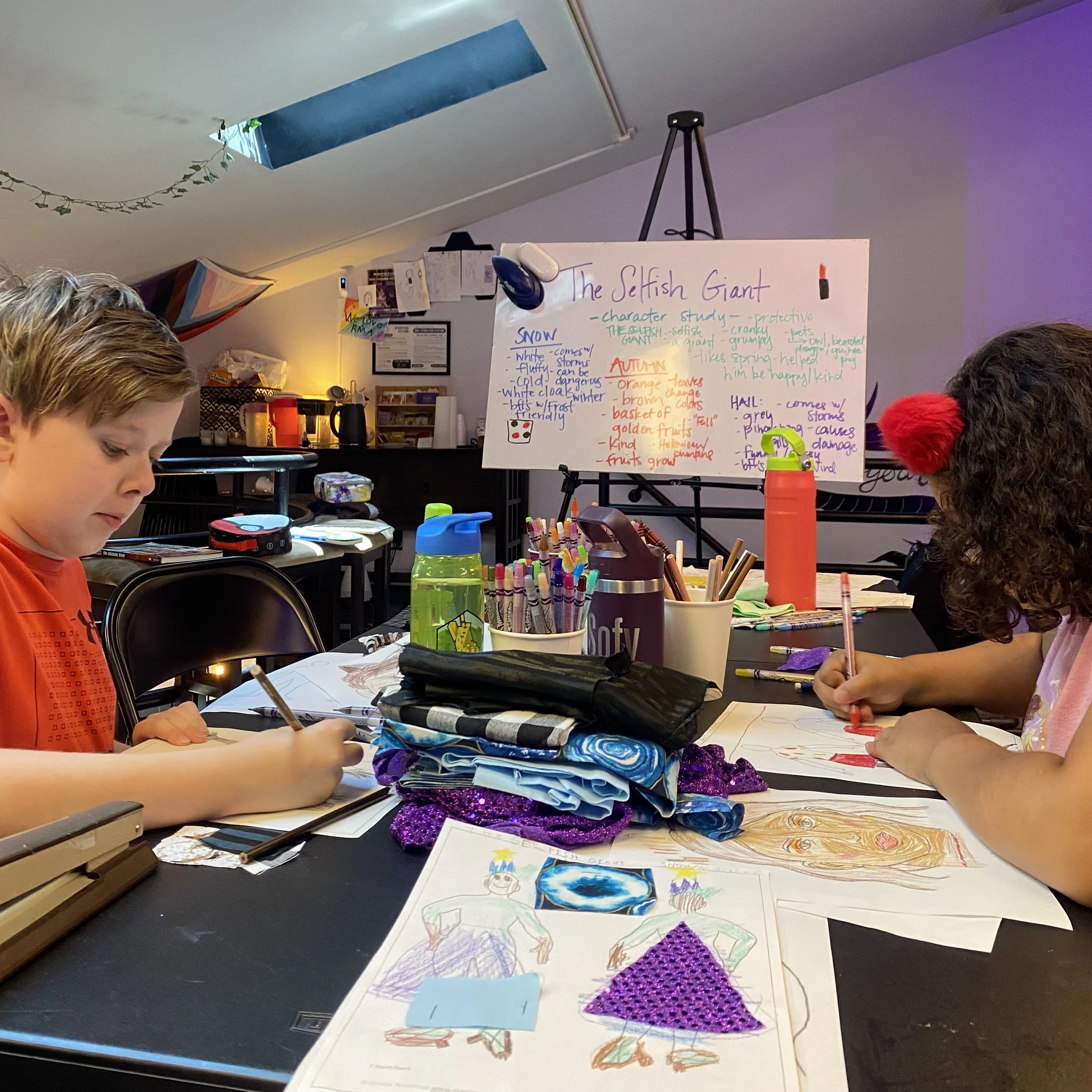 Children in a classroom working on art projects with drawings, glitter, and markers on tables, and a whiteboard with notes about 'The Selfish Giant' in the background.
