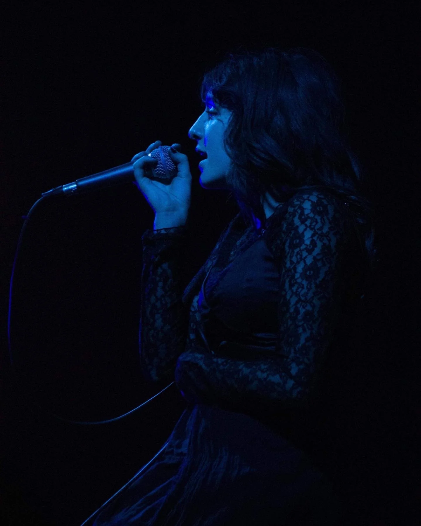 A woman singing into a microphone on stage, illuminated by blue lighting, with dark background.