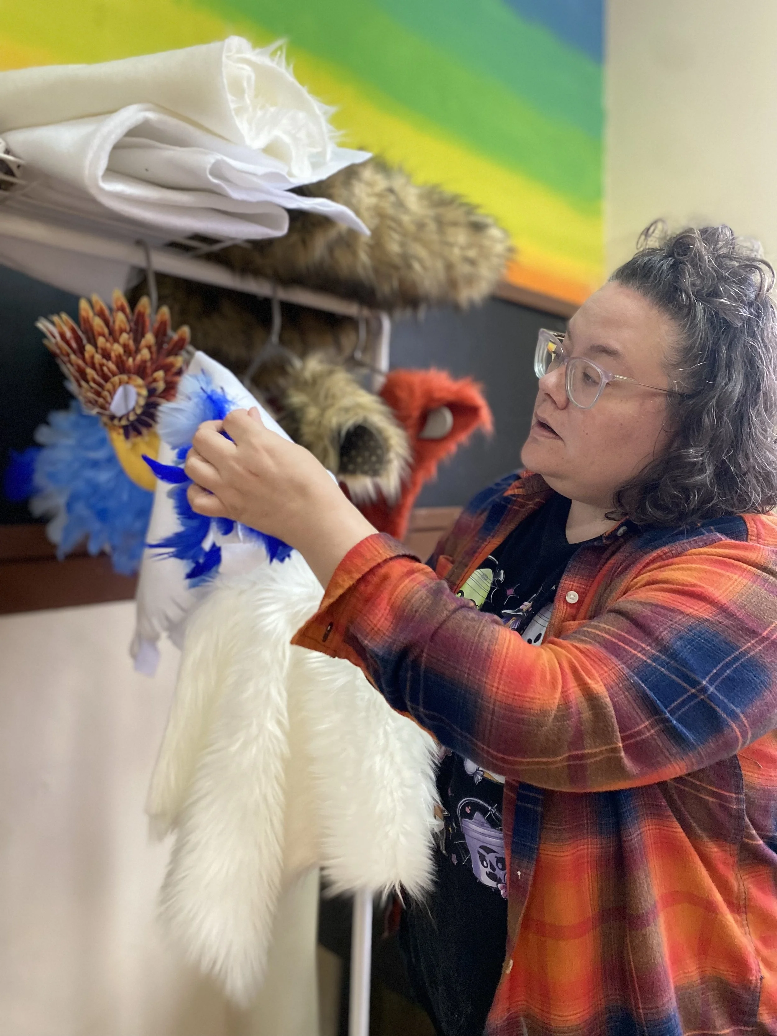 A woman with curly hair and glasses is adjusting a white costume that appears to be a mascot or animal costume, with a blurred background featuring a colorful rainbow mural.