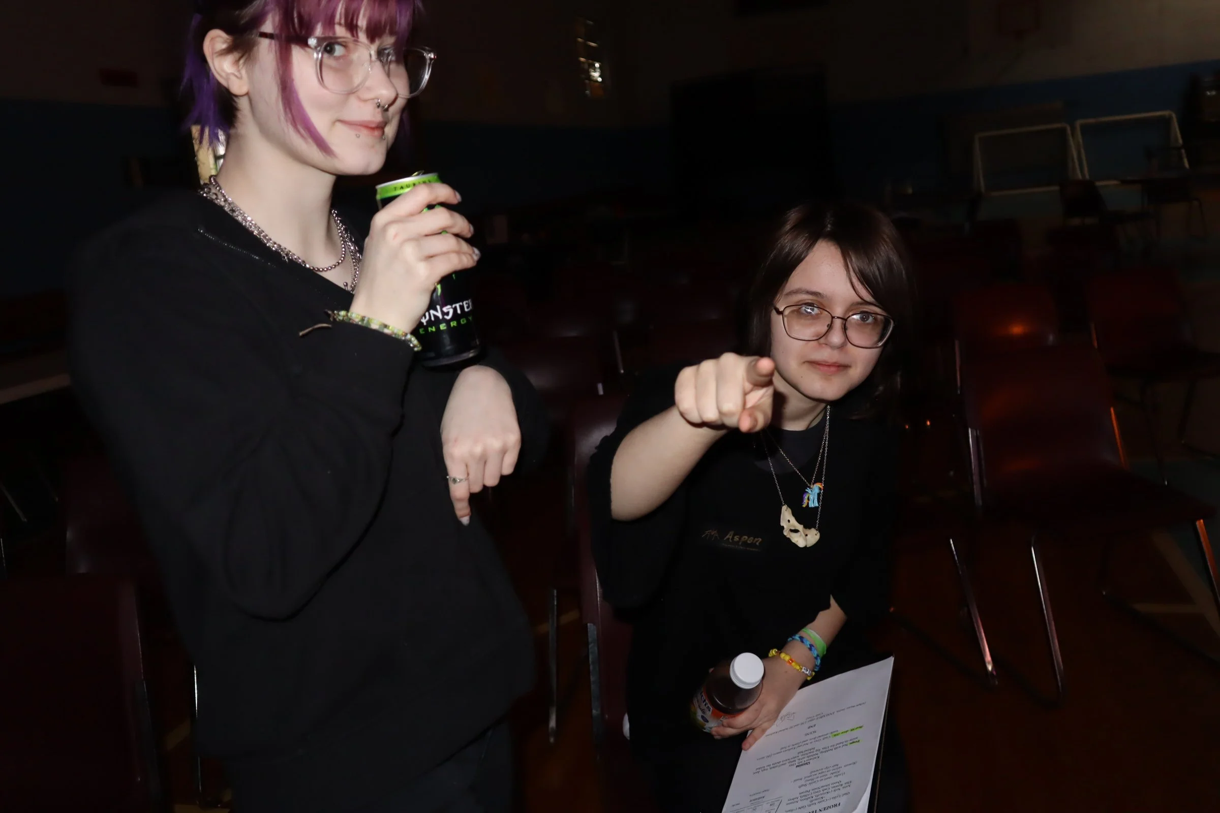Two young women sitting in a dark room with chairs in the background. The woman on the left has purple-dyed hair, glasses, and multiple facial piercings. She is holding a can of Monster energy drink. The woman on the right has long dark hair, glasses, and is pointing towards the camera while holding a bottle and some papers.