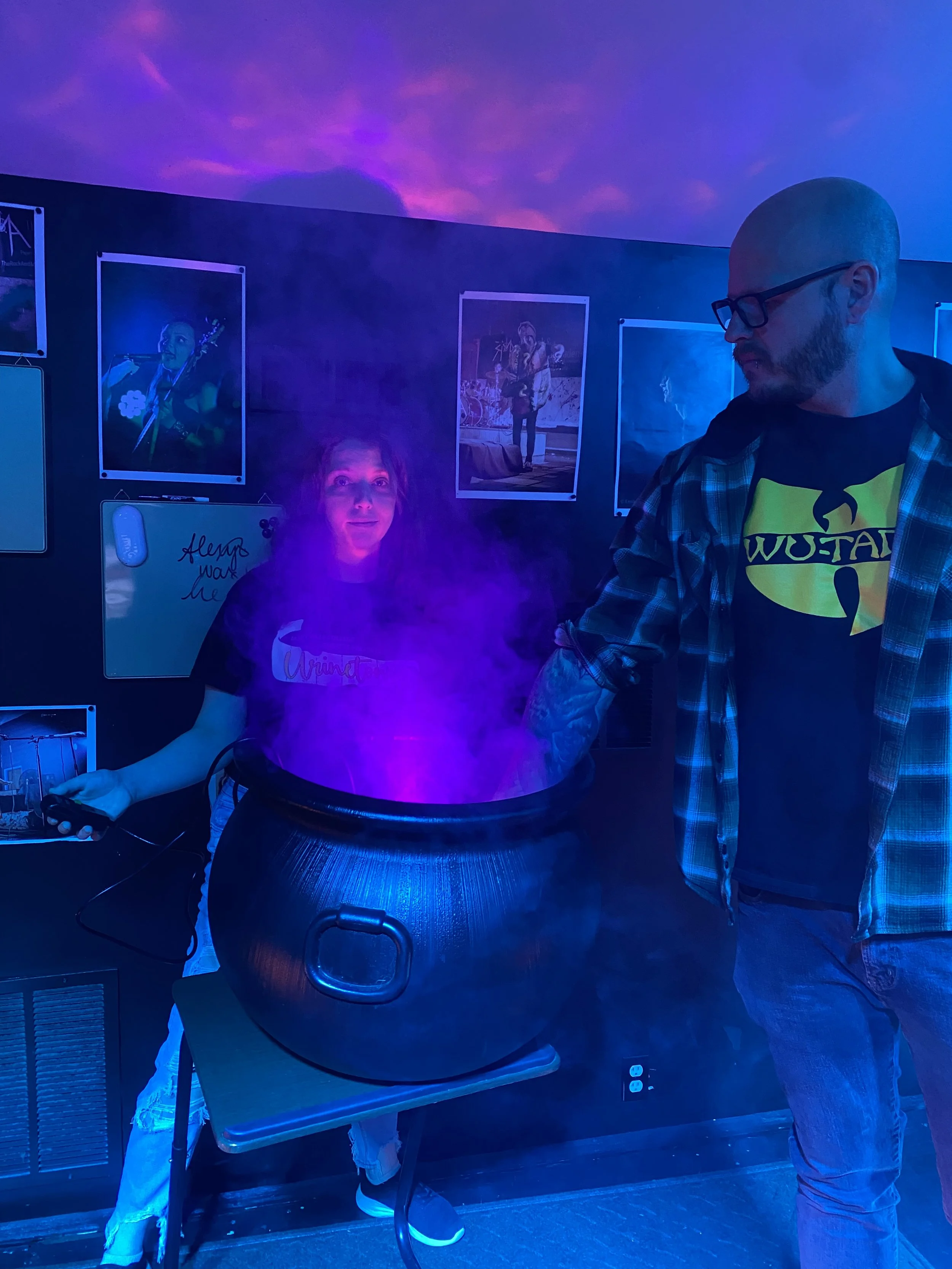 Two people in a dark room with blue and purple lighting, one person is holding a device, and the other is standing near a bubbling cauldron emitting purple smoke, with posters on the wall behind them.