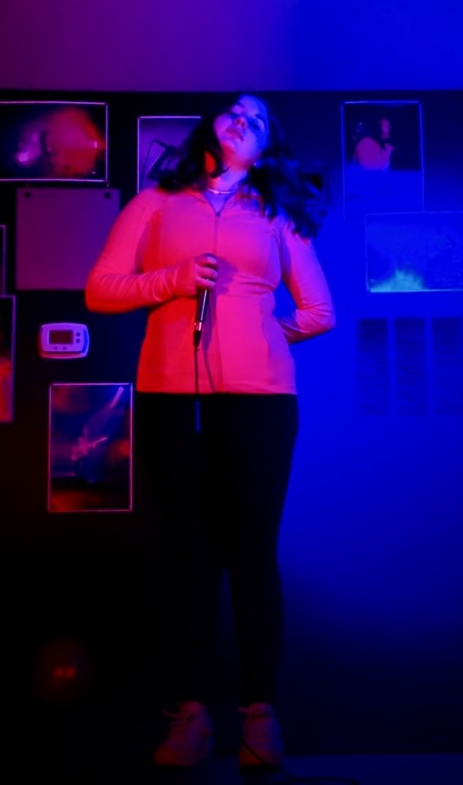 Woman holding a microphone in a dimly lit room with colorful neon lighting, standing in front of a wall decorated with framed photos or artwork.