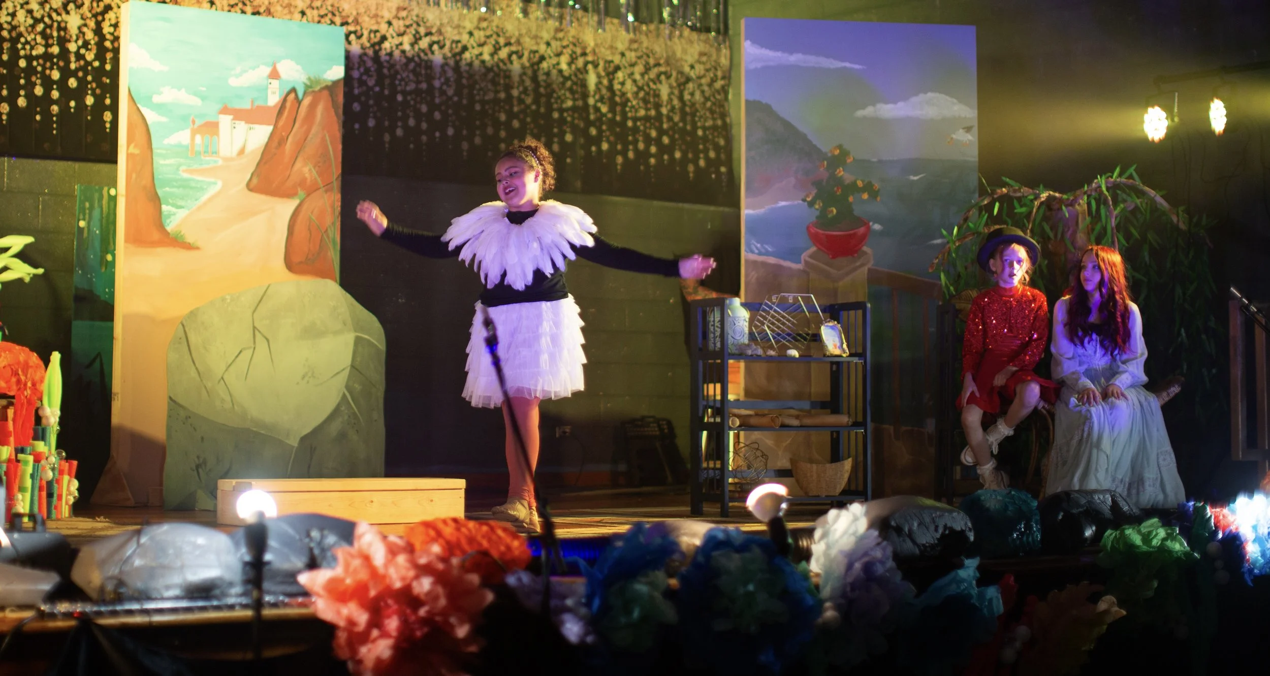 A stage performance with three young girls; one in the foreground wearing a black dress with white feathered collar and skirt, smiling with arms outstretched. Two girls are sitting to the right, one in a red sparkly dress and the other in a dress. The background has painted scenery of mountains, trees, and ocean, with colorful props and decorations on stage.