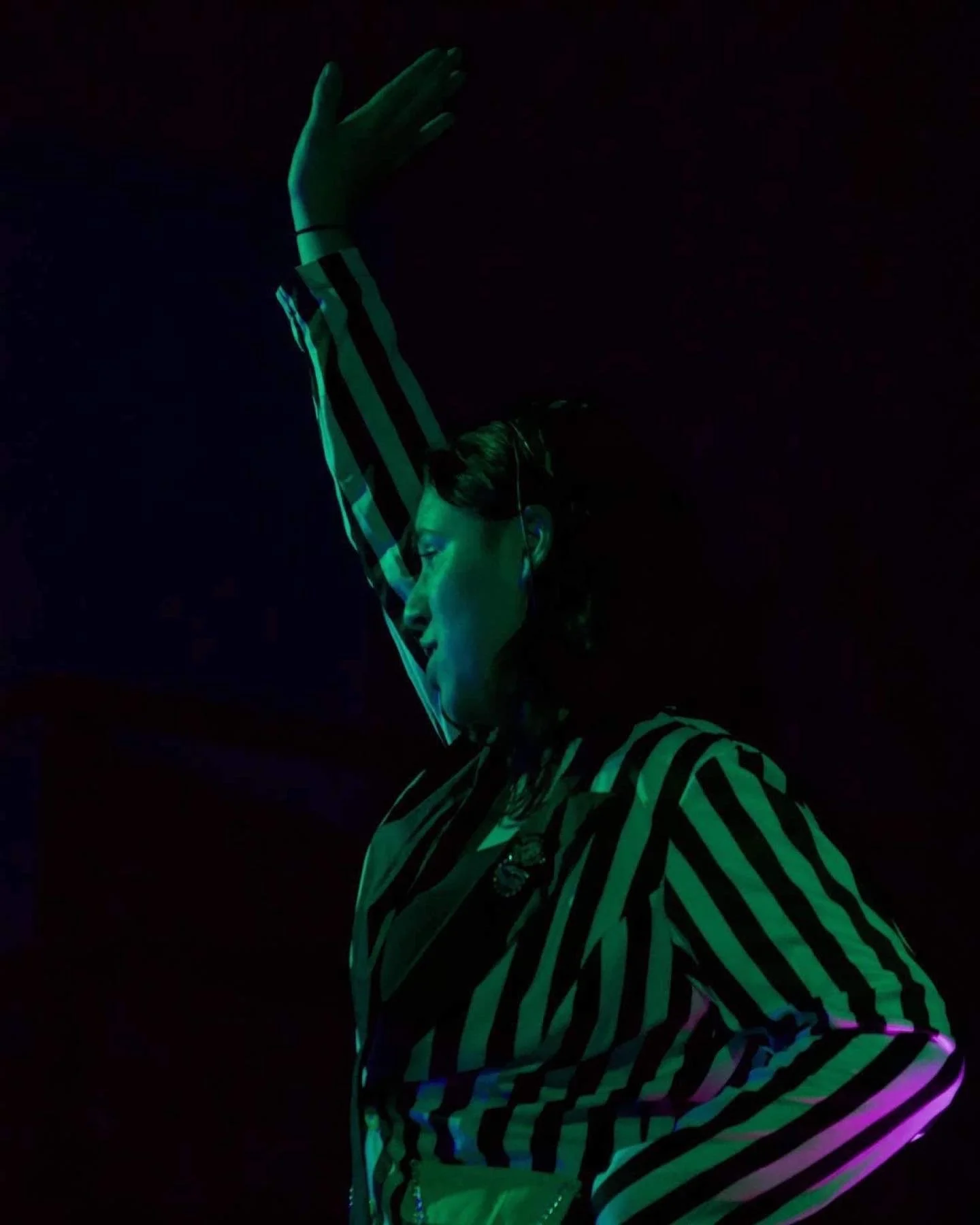Person with closed eyes, wearing a black and white striped outfit, raising one arm above their head in a dark setting with colorful lighting.