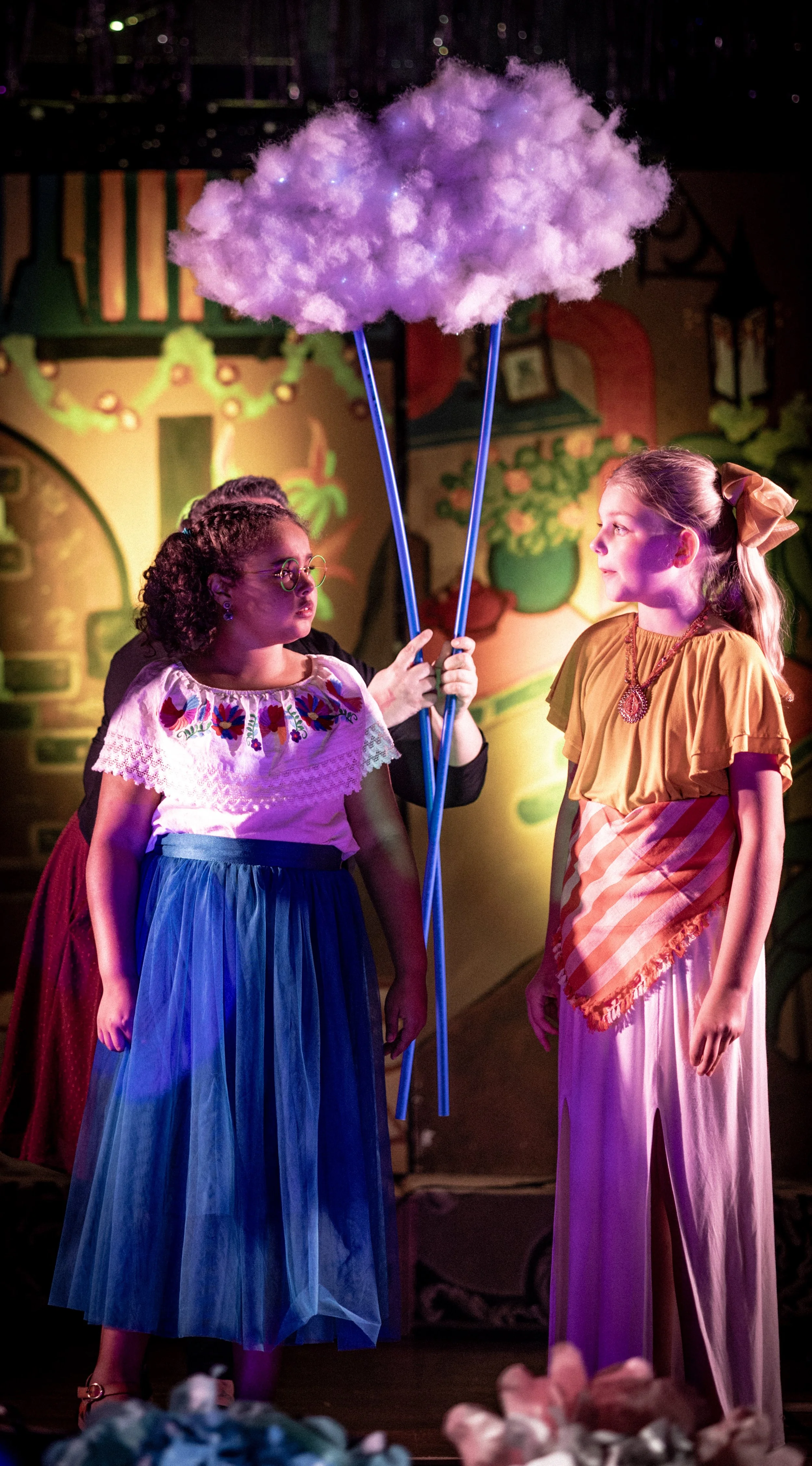 Two young girls in colorful costumes stand on stage under a purple cotton candy prop held by an adult. The background features a vibrant, whimsical set design with bright colors, large decorative elements, and theatrical lighting.