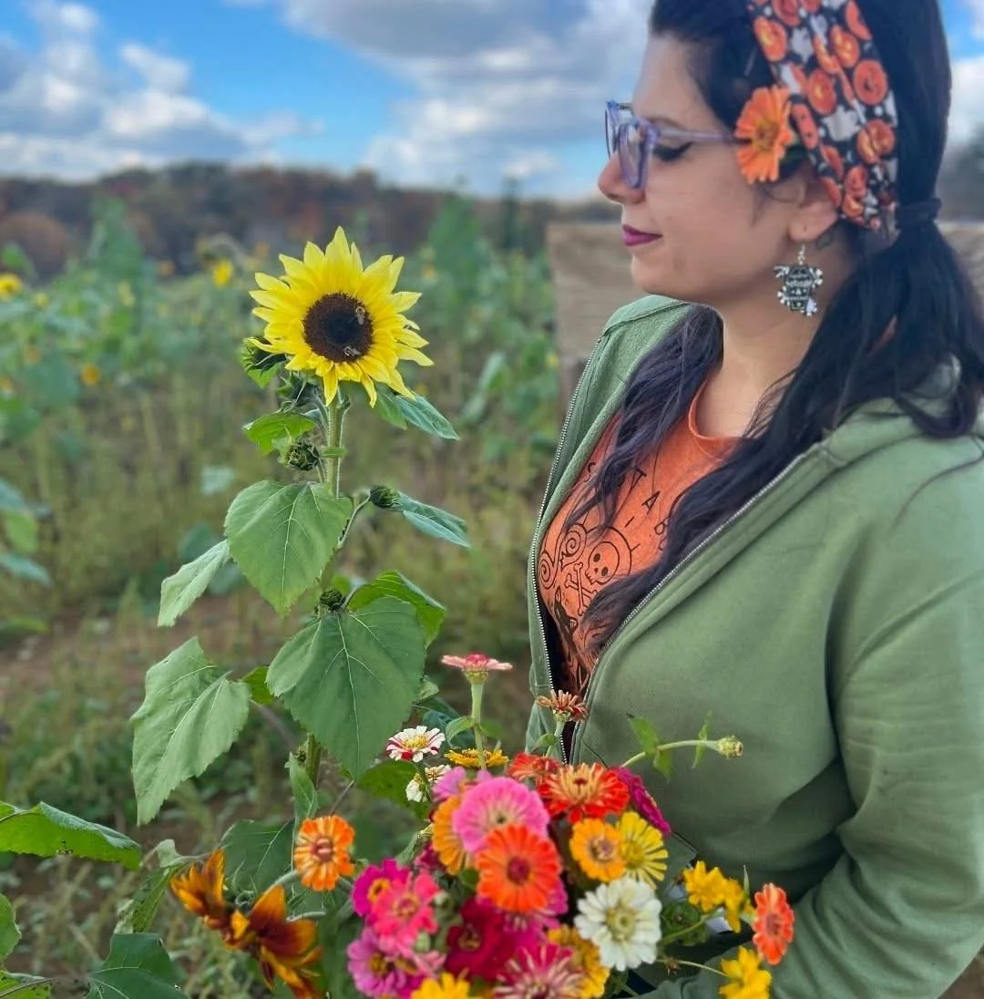 A woman wearing glasses, a floral headband, and earrings stands in a field of sunflowers and colorful flowers, holding a bouquet of flowers, with a partly cloudy sky in the background.