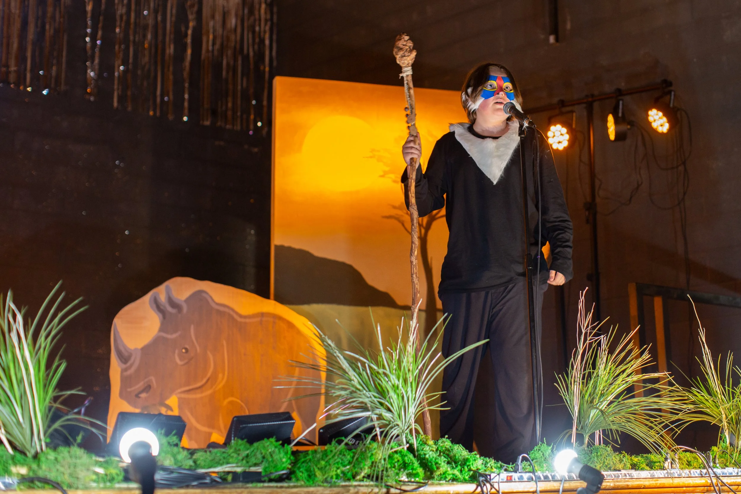 Person dressed as a Native American, wearing a face mask and feathers, standing behind a microphone on stage. The stage has a backdrop depicting a sunset with a tree silhouette. There are plants and lighting on the stage floor.
