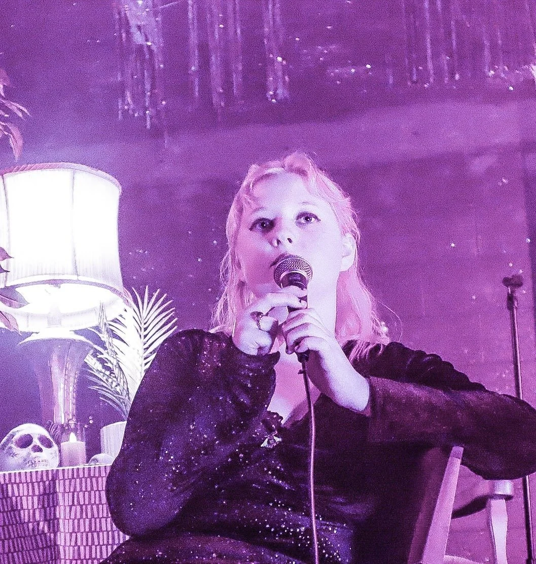 A woman with pink hair holding a microphone, sitting in a room with purple ambient lighting, with a lamp, plant, skull, and candles in the background.