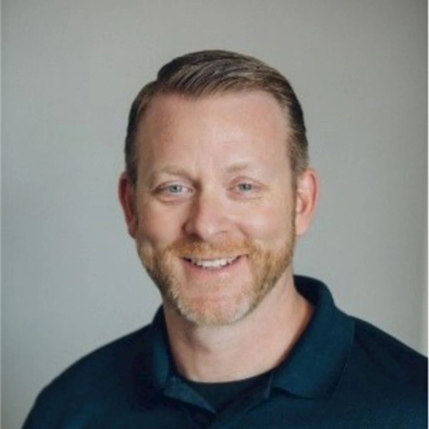 Headshot of a smiling man with short brown hair, light beard, wearing a dark collared shirt, against a plain gray background.