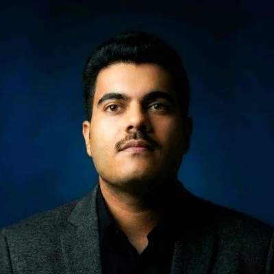 A portrait of a man with dark hair and a serious expression, wearing a dark blazer against a dark blue background.
