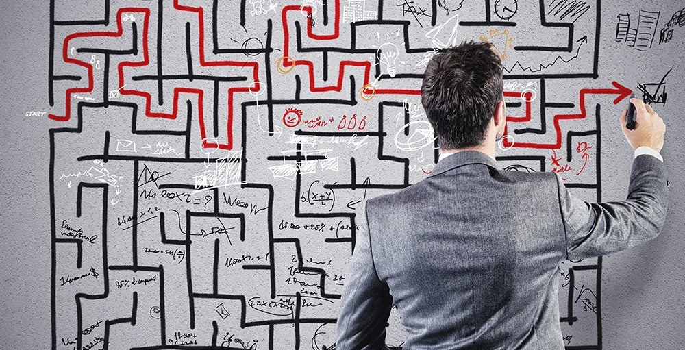 Man in gray suit drawing a red line on a complex maze diagram on a whiteboard filled with various symbols, notes, and sketches.