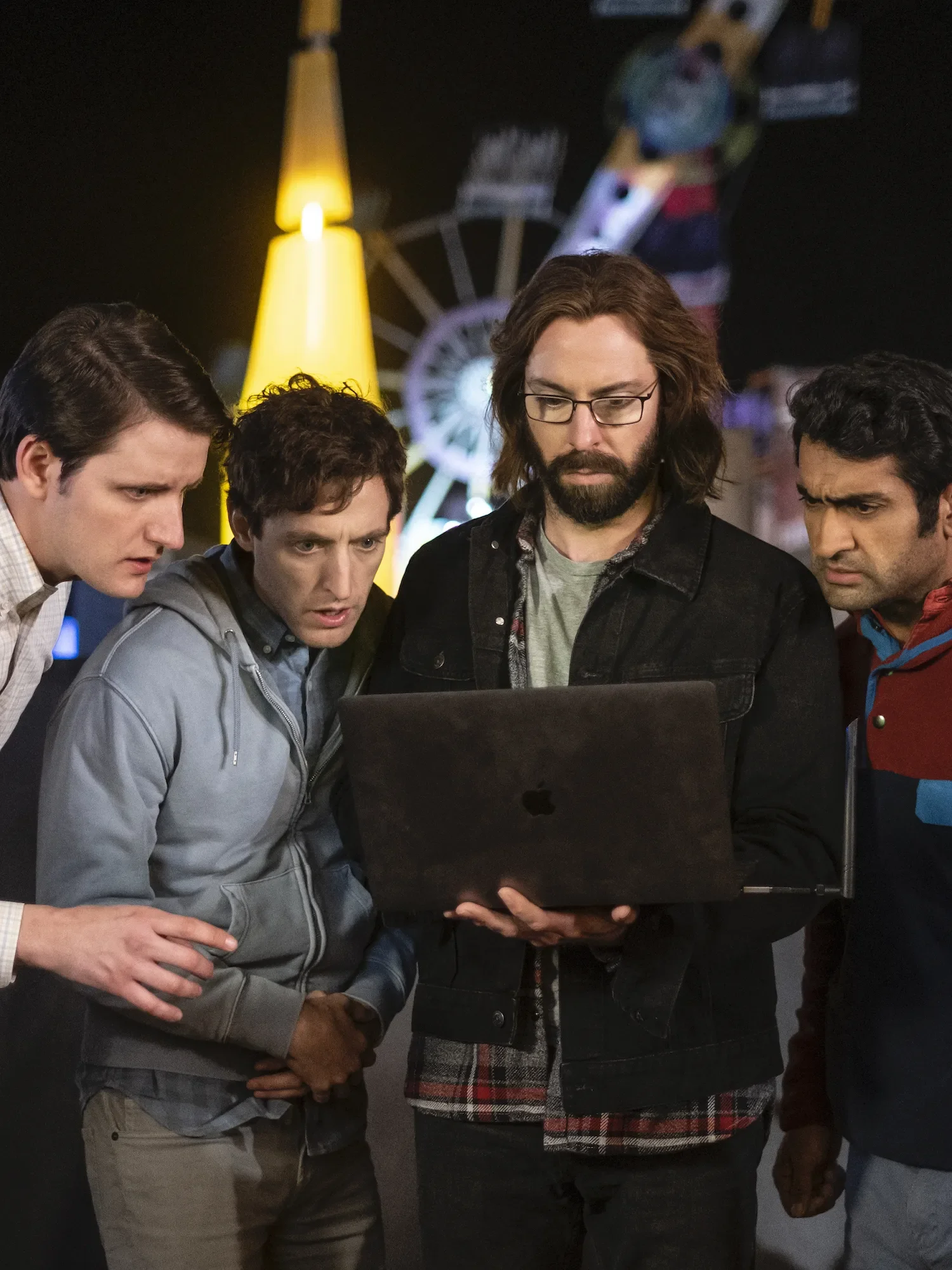 Four men standing closely together looking at a laptop screen in front of a Ferris wheel and a yellow lamp at night.