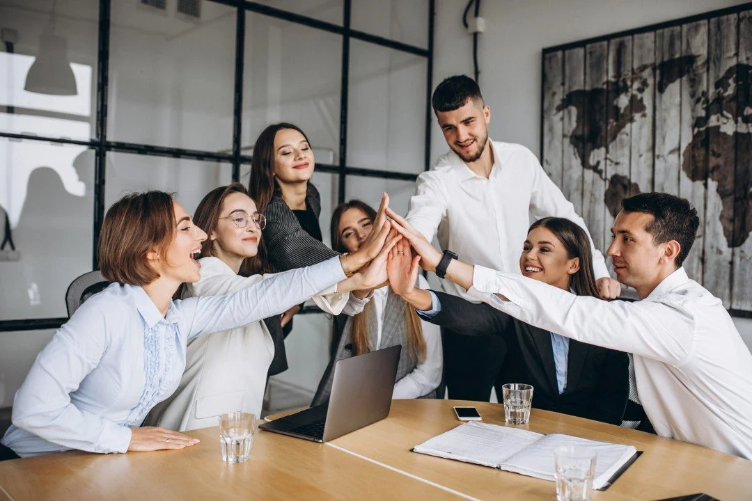 A diverse group of coworkers in a business meeting giving a group high five around a table.