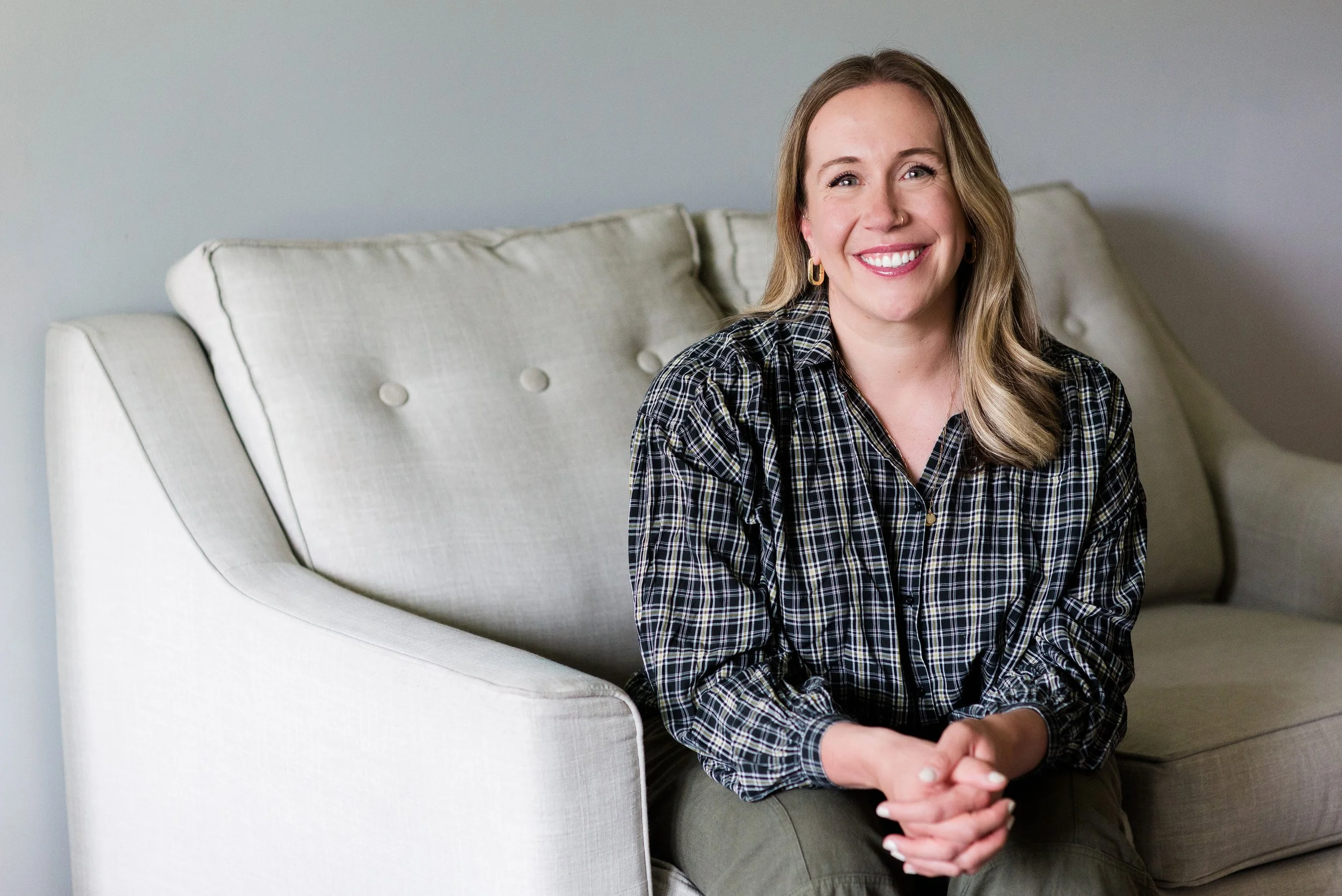 A woman with long, wavy blond hair and gold hoop earrings is sitting on a light-colored sofa, smiling and looking at the camera. She is wearing a black and white plaid shirt.