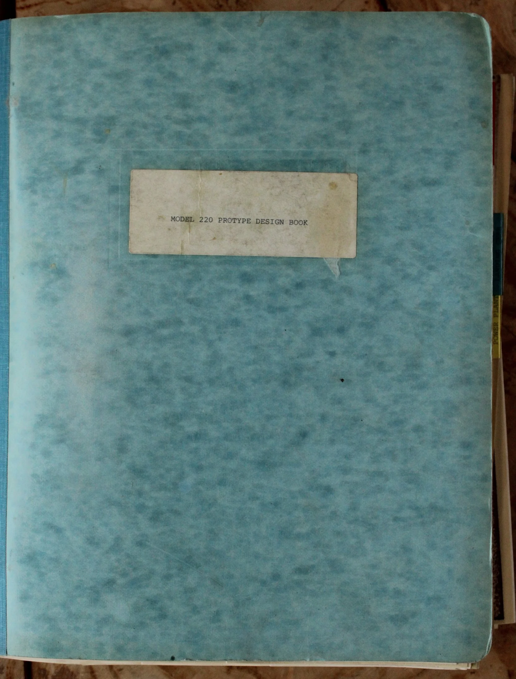 A light blue, spiral-bound notebook with a white label in the center that reads "Model 220 Prototype Design Book."