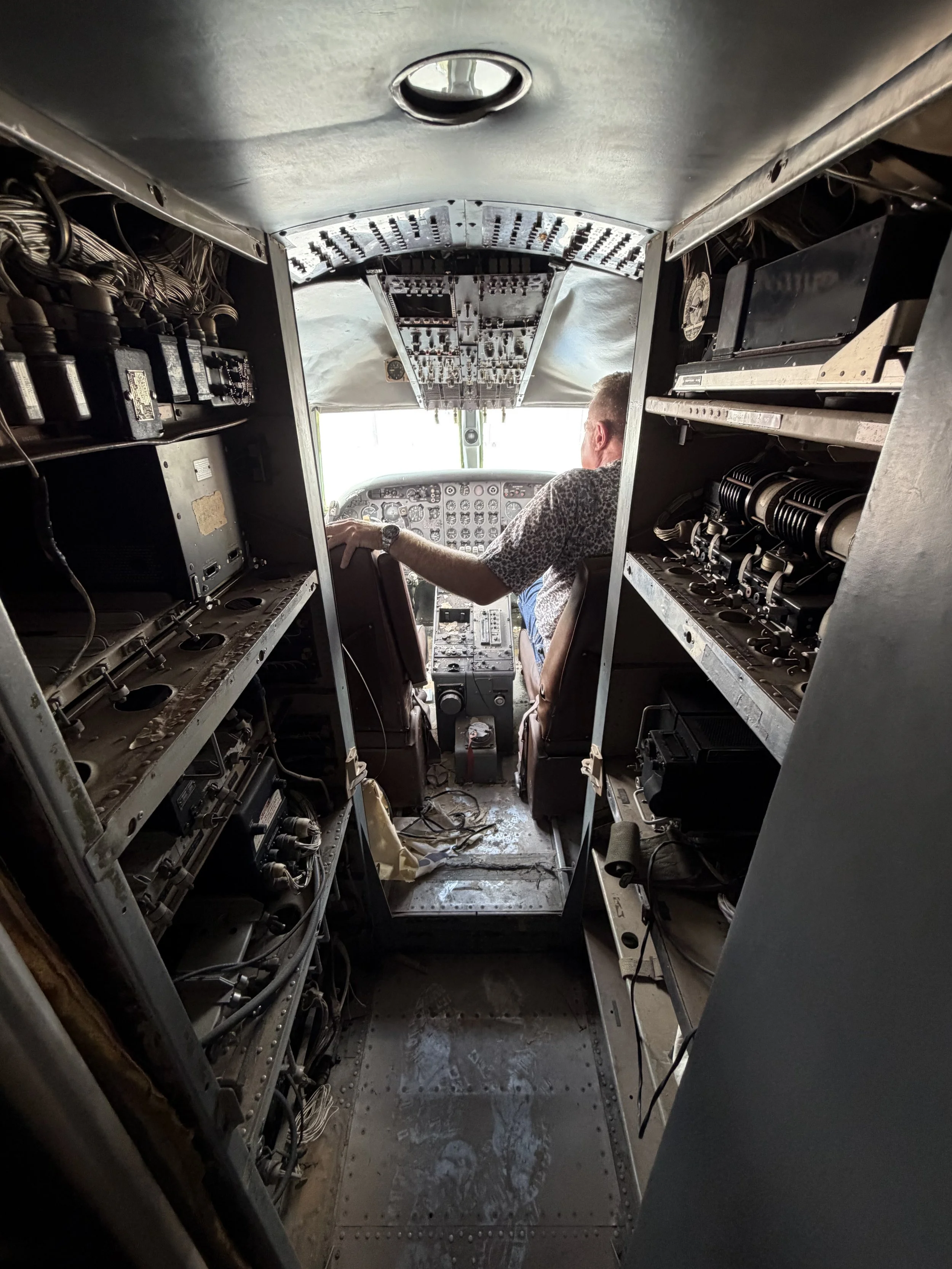 View inside the cockpit of an aircraft showing the pilot seated and various control panels and electronic equipment surrounding him.