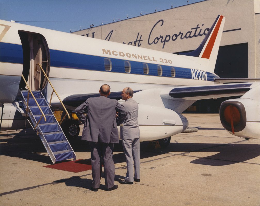 Two men in suits stand near a vintage McDonnell 220 private jet on an airport tarmac, conversing outside the aircraft with stairs leading up to the open door.