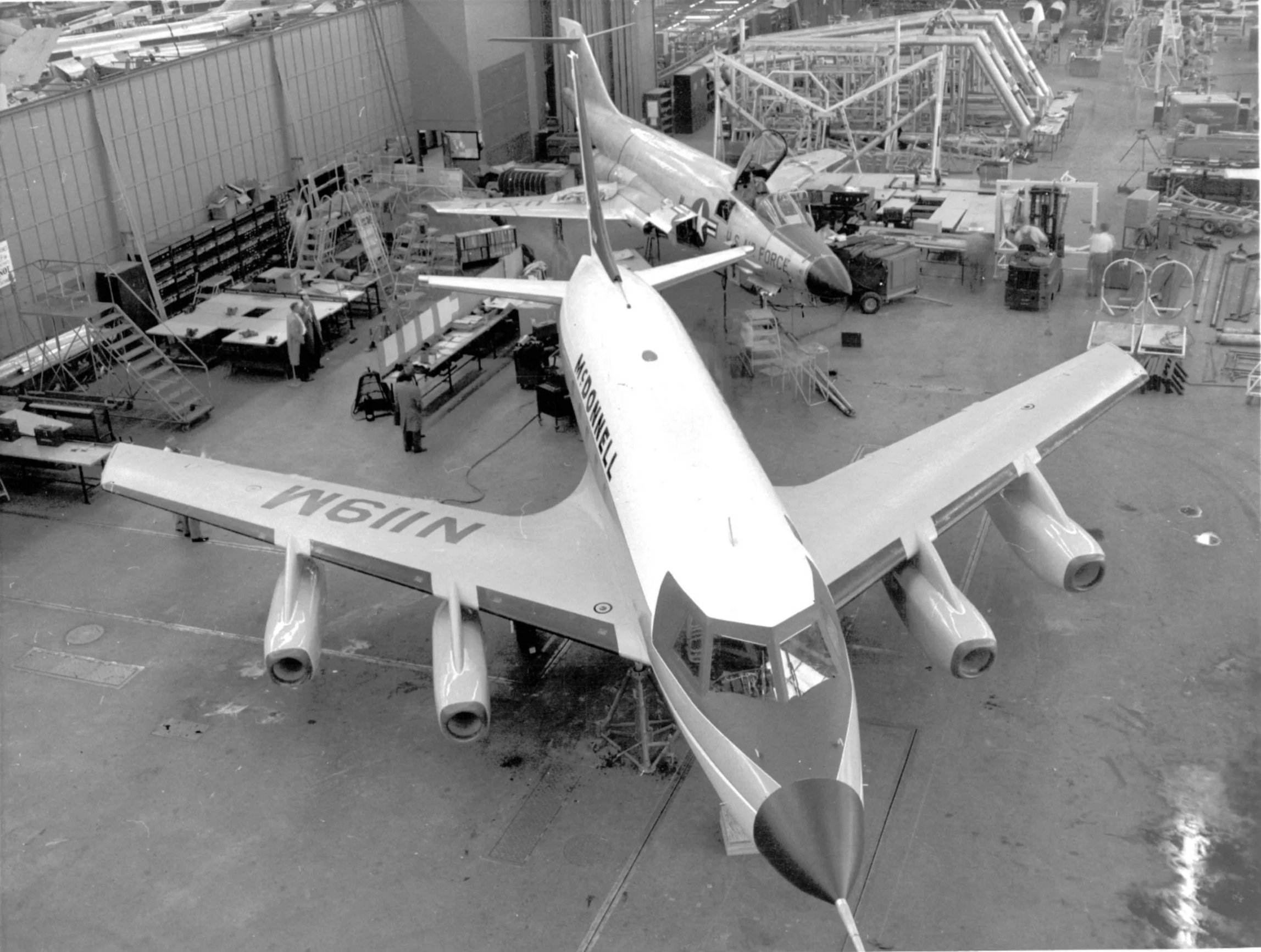 Black and white photo of an aircraft inside a manufacturing or maintenance facility with workers and equipment around.