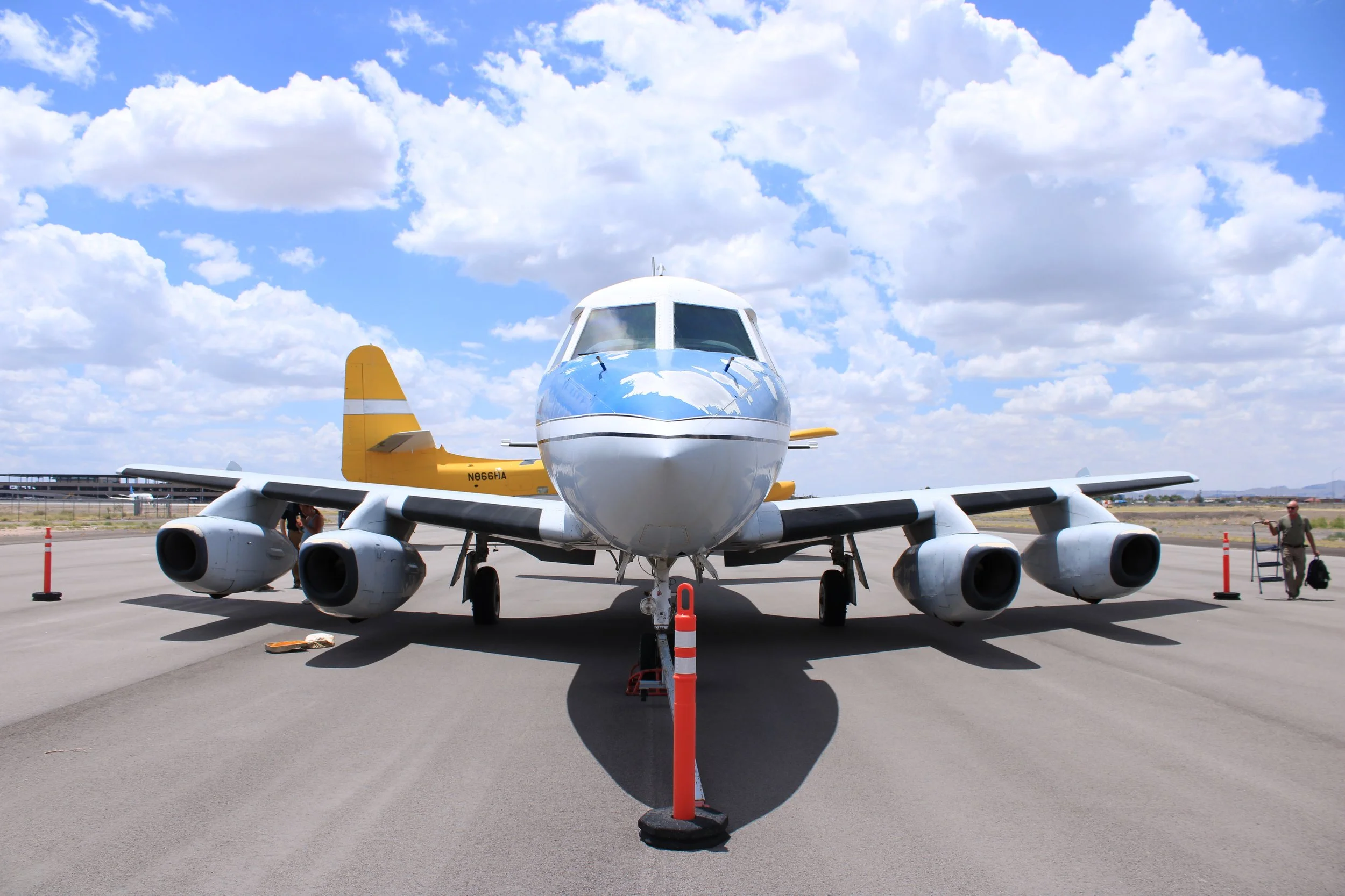 A small white and yellow airplane parked on a tarmac under a partly cloudy sky, with orange cones and a few people nearby.