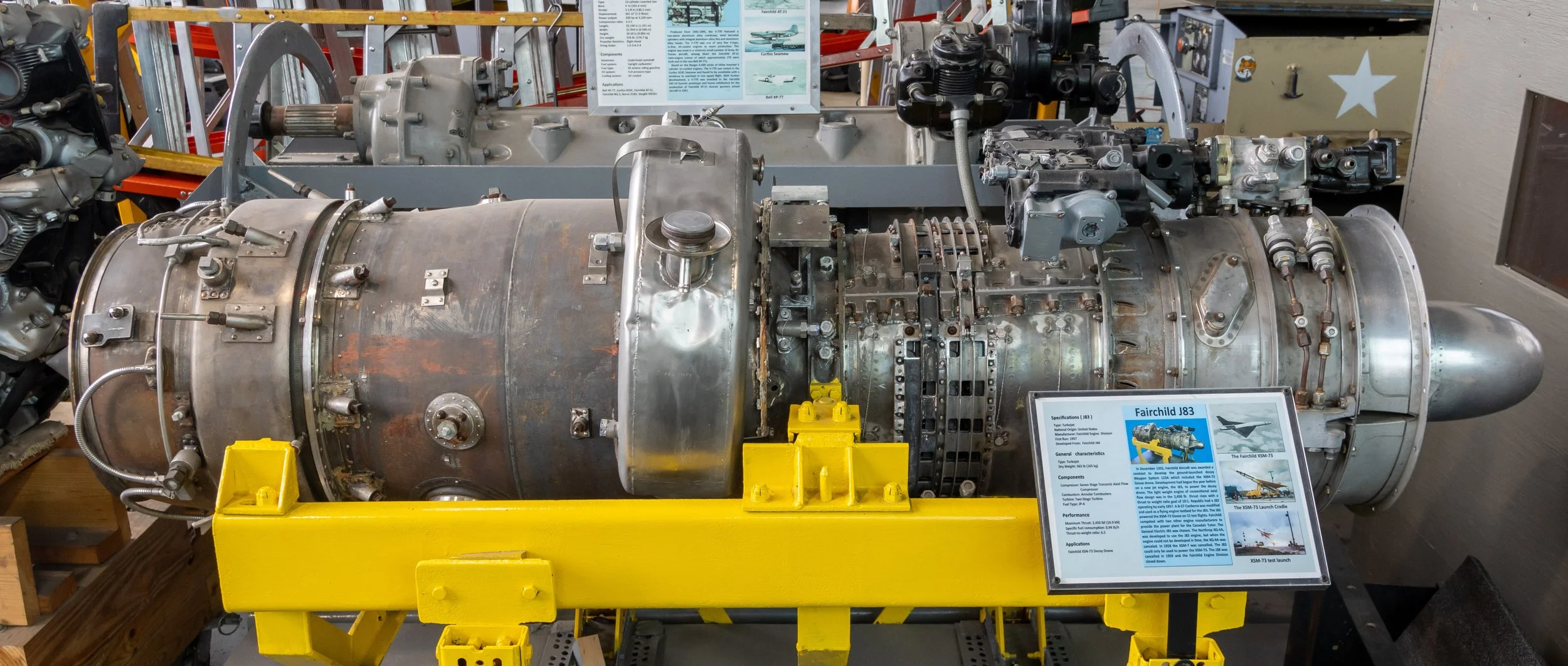 Aircraft engine labeled 'Fairchild J83' on display with informational signs behind it, surrounded by other aircraft parts and equipment in a museum setting.