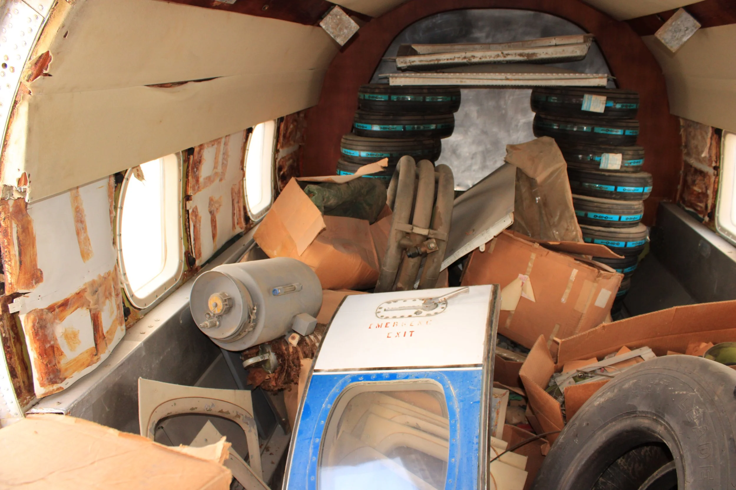 Disorganized interior space with tires, cardboard boxes, and equipment, including a white machine with a label that reads 'Emergency Exit' and walls with peeling paint and exposed insulation.