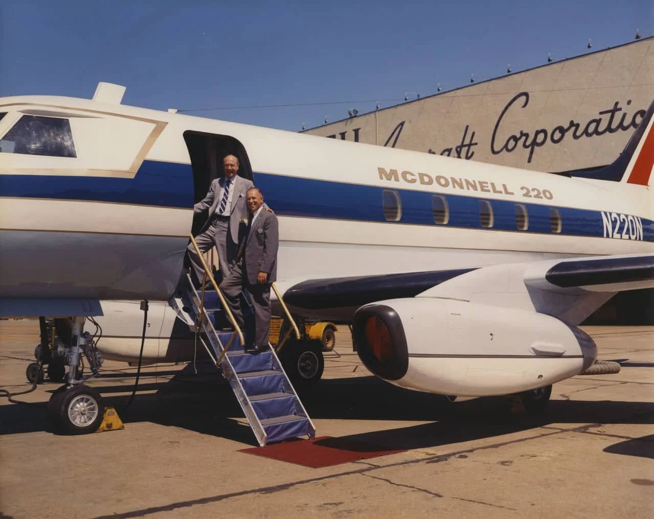 Two men in suits standing on a staircase leading to the door of a large white and blue airplane with the registration N220N, labeled 'McDonnell 220.' They are on an airport tarmac, and a building in the background has the words 'East Corporation' visible.