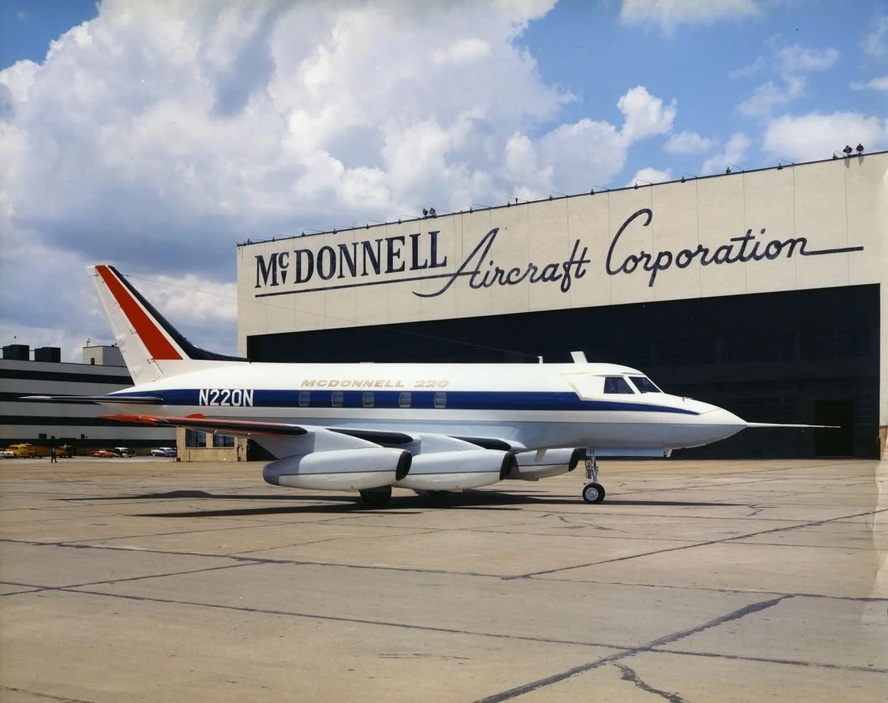 A small vintage white aircraft with blue and red accents parked on the tarmac outside of a large aircraft hangar. The hangar has 'McDonnell Aircraft Corporation' written on it. The sky is partly cloudy with a few clouds visible.