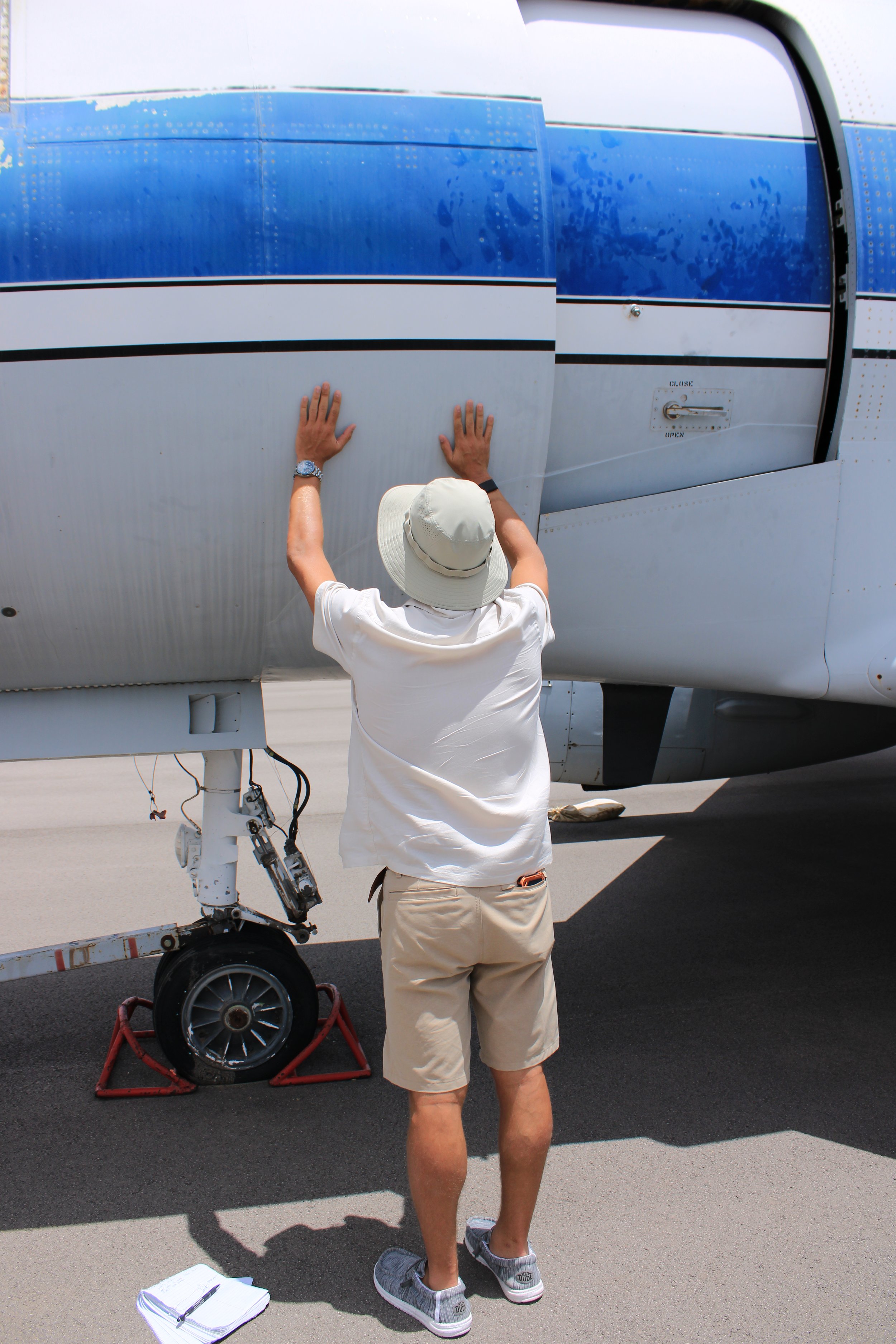 A person wearing a white wide-brimmed hat, beige shorts, and gray sneakers, working on the landing gear of an airplane. The person is standing on the tarmac, with tools and paperwork on the ground nearby. The airplane's blue and white exterior is visible.